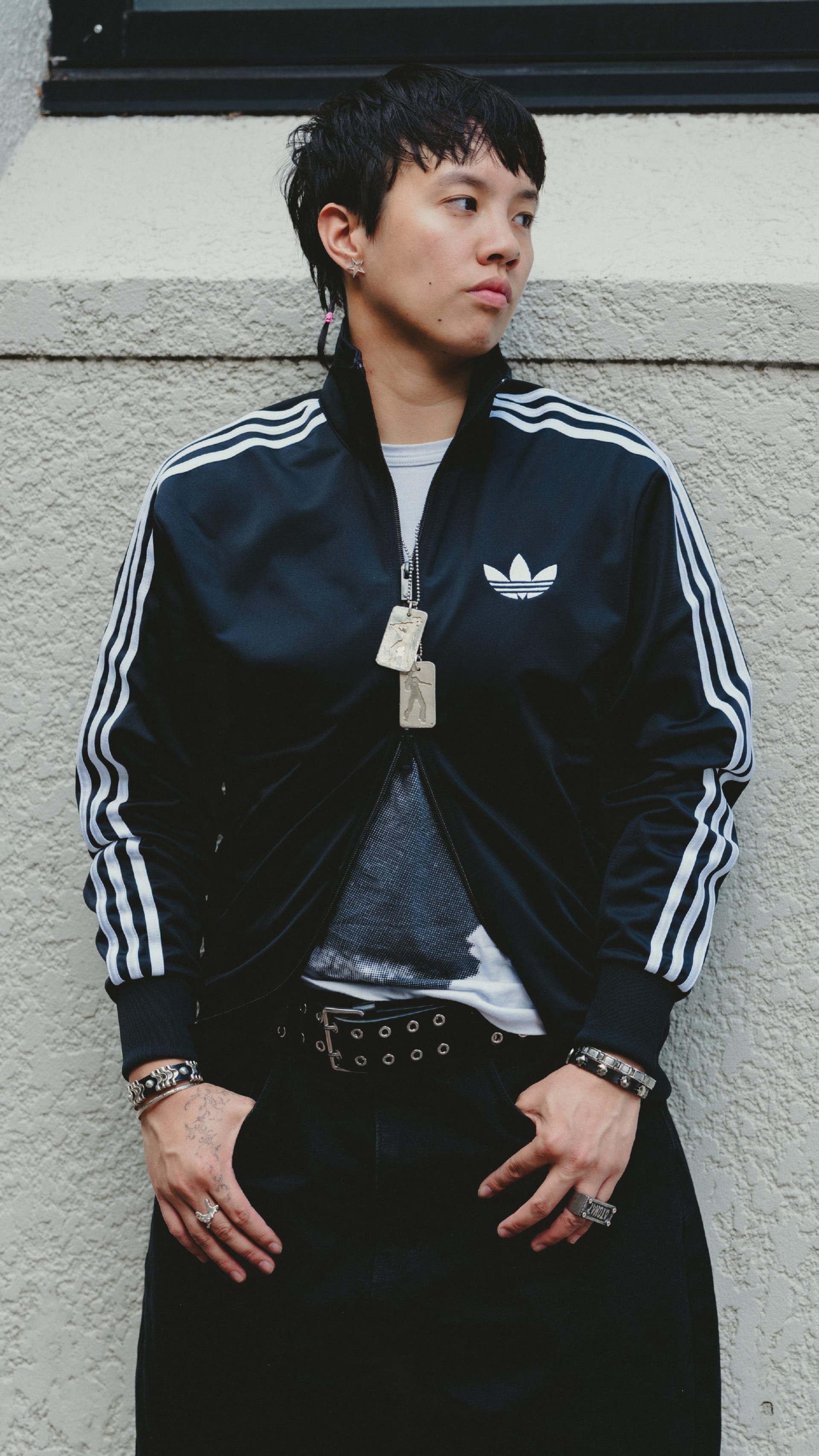 A young person wearing a black Adidas jacket with white stripes stands in front of a white wall.