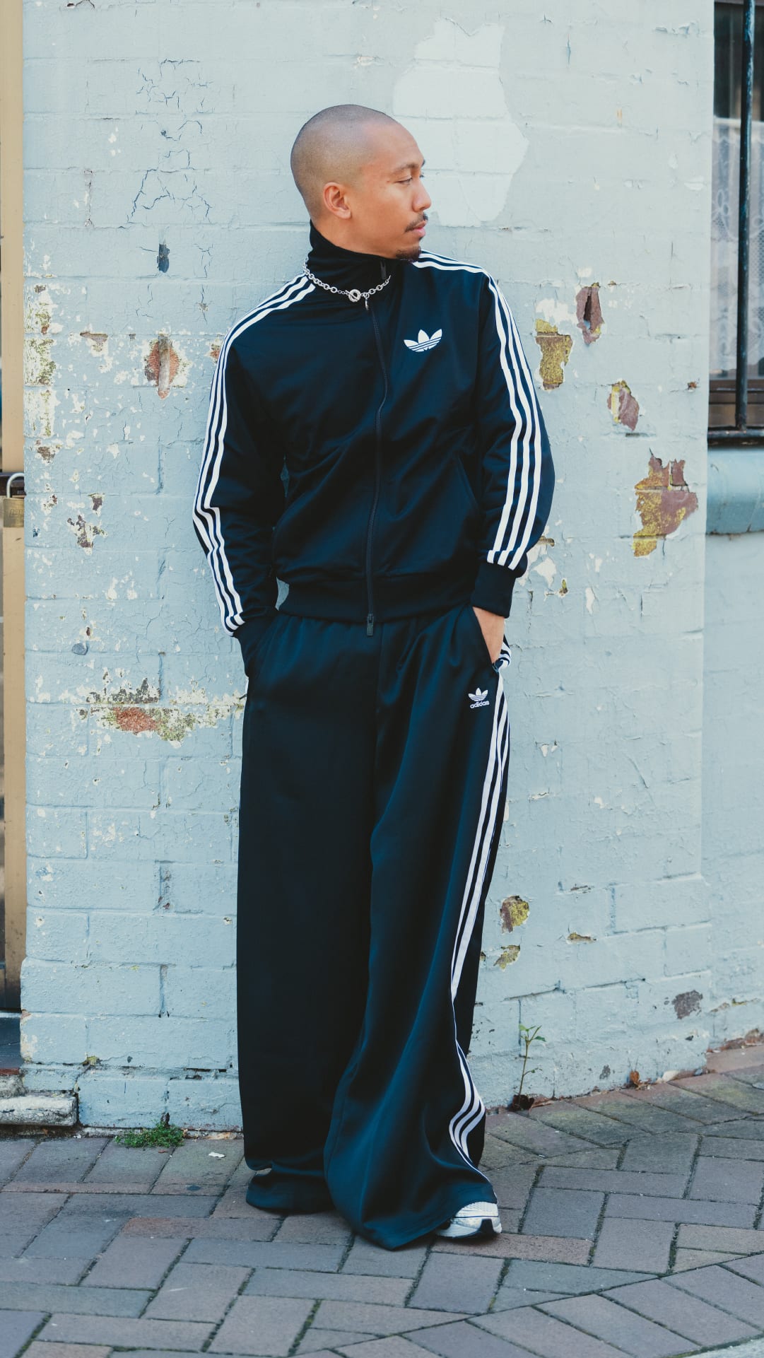 A person wearing a black and white tracksuit stands in front of a dilapidated brick wall.