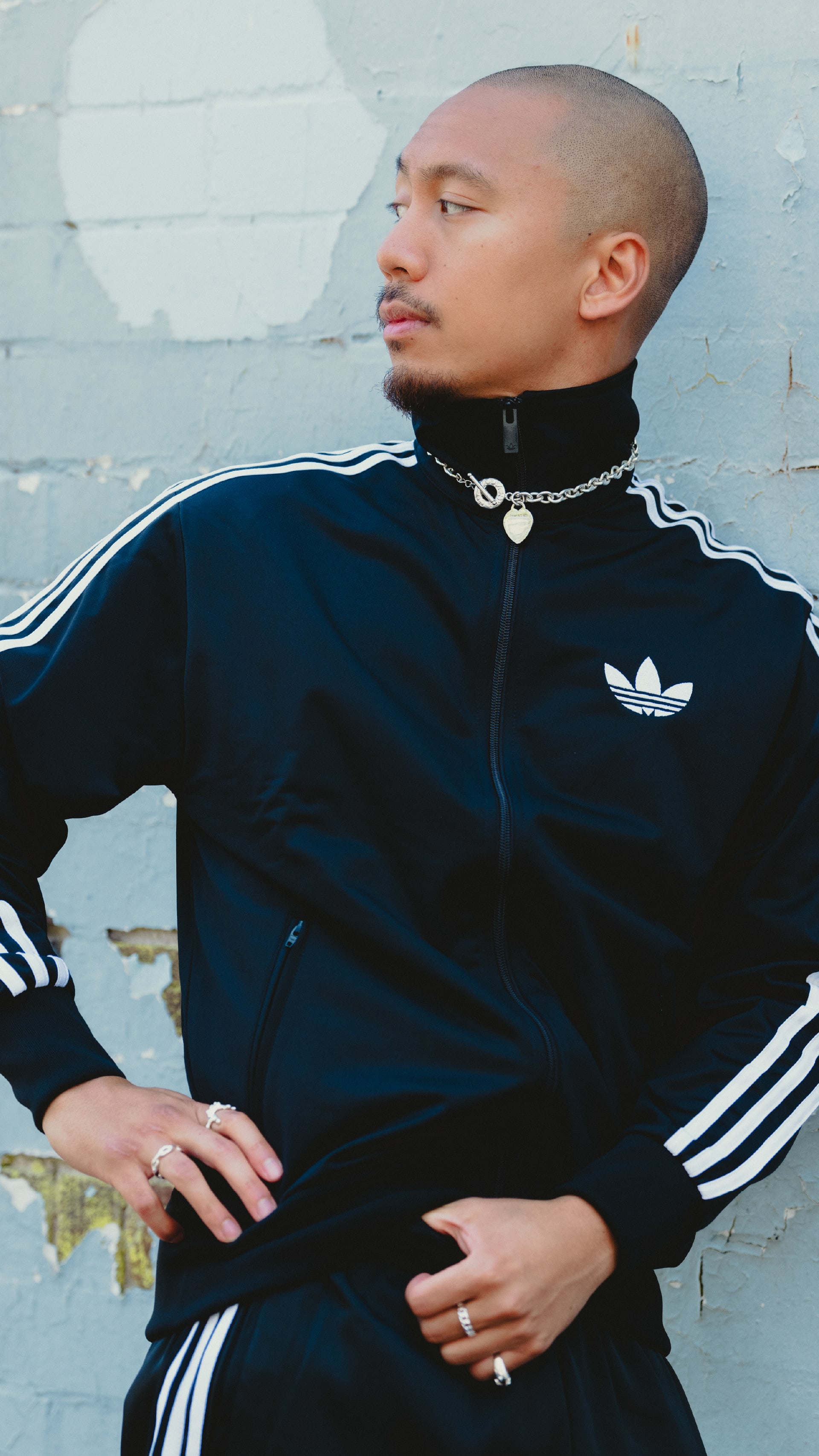 A person wearing a black Adidas tracksuit jacket stands in front of a white wall, with a serious expression on their face.