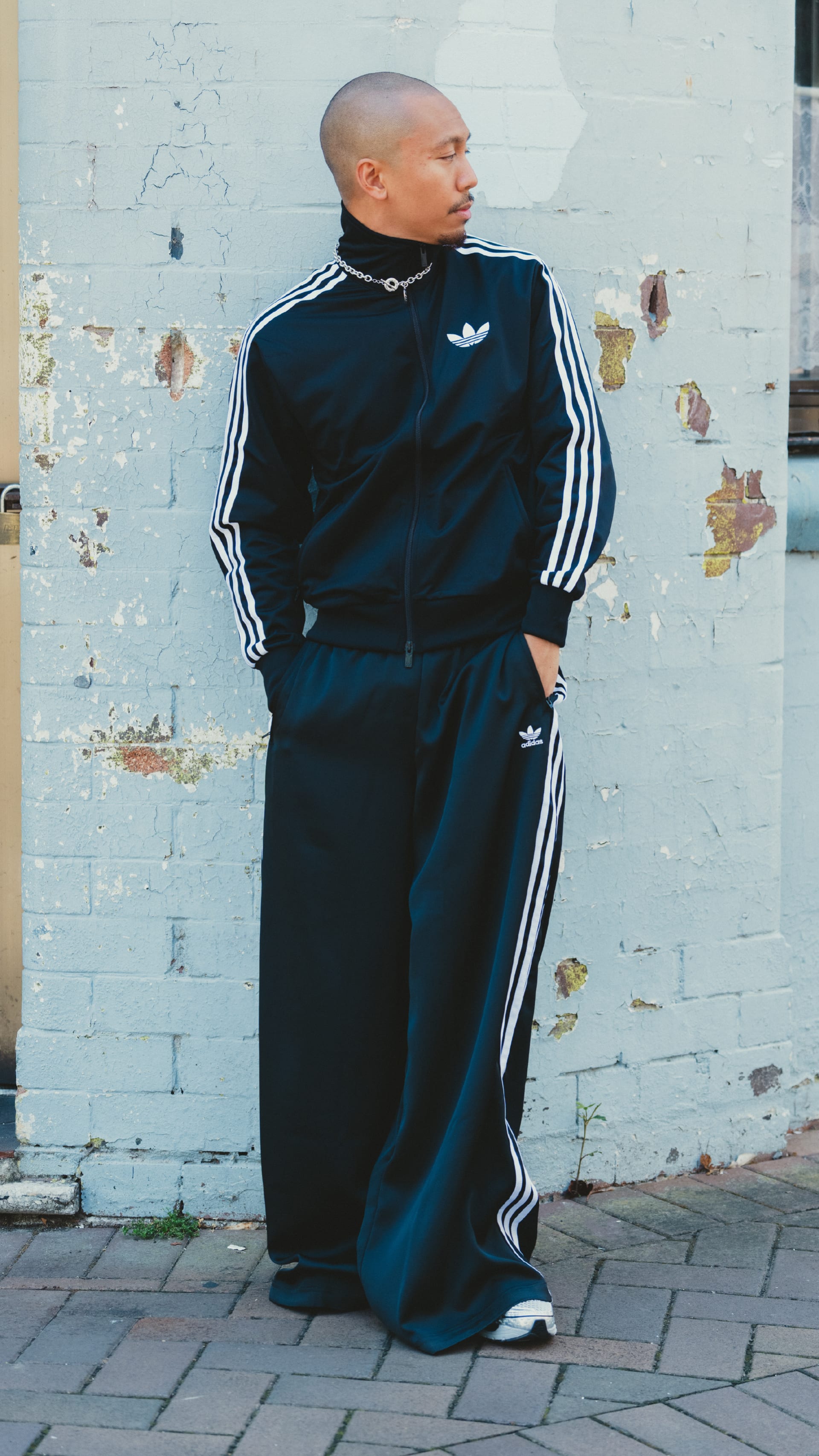 A person wearing a black tracksuit with white stripes stands in front of a dilapidated wall, their expression pensive.