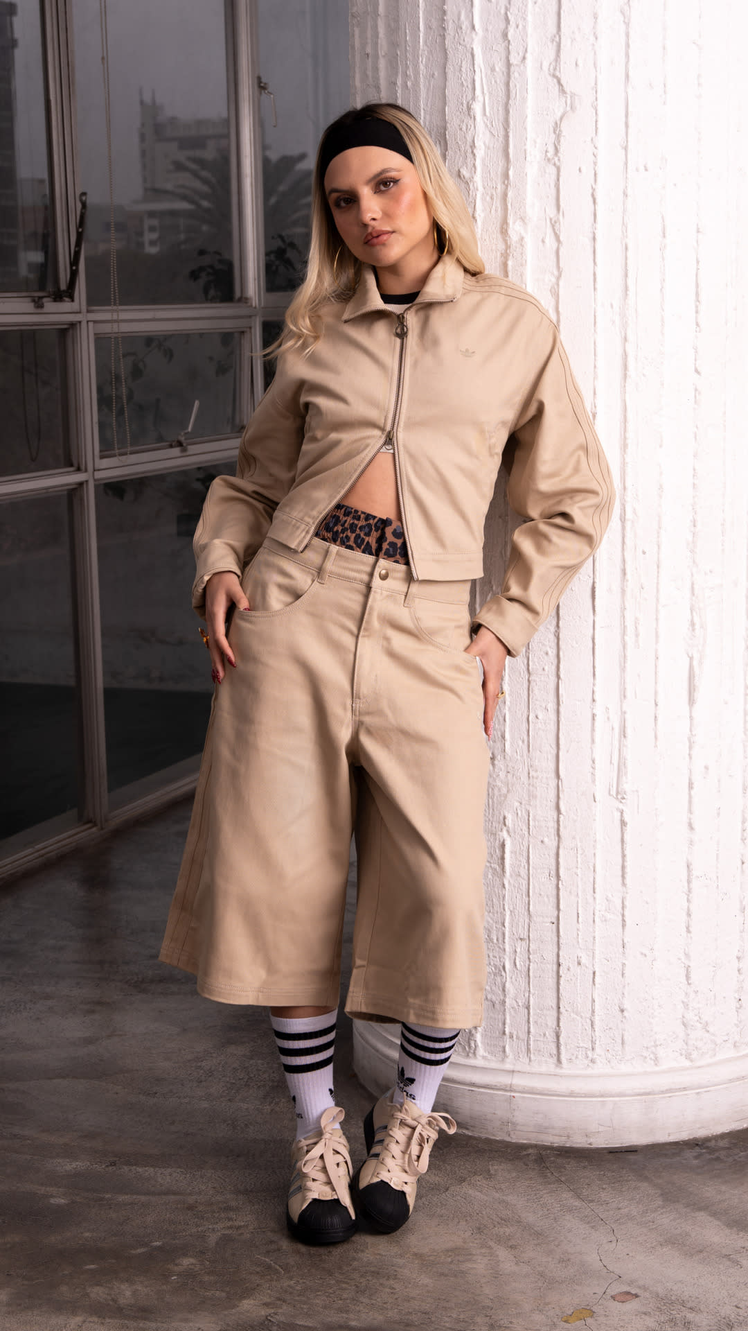 A person wearing a beige hooded jacket and pants stands in front of a window, with a brick wall visible in the background.