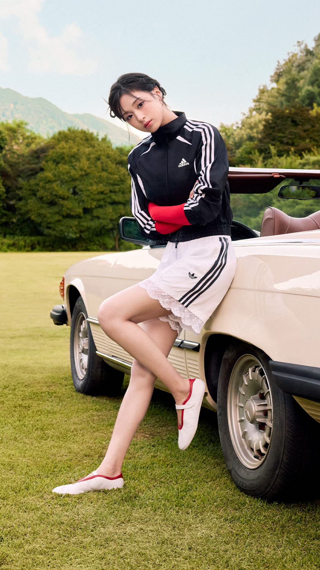 A person in a black and white athletic outfit is sitting on the edge of a classic car parked on a grassy field, with mountains visible in the background.