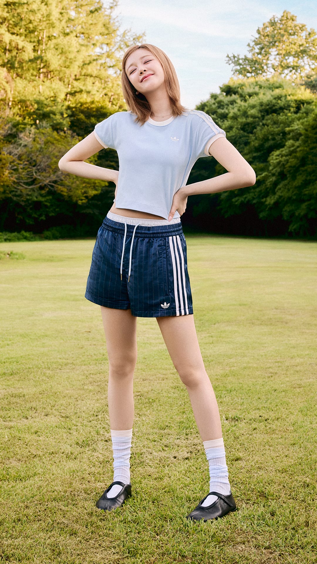 A young woman stands in a grassy field, wearing a white t-shirt and navy blue shorts with white stripes, her arms raised as she enjoys the outdoors surrounded by trees.