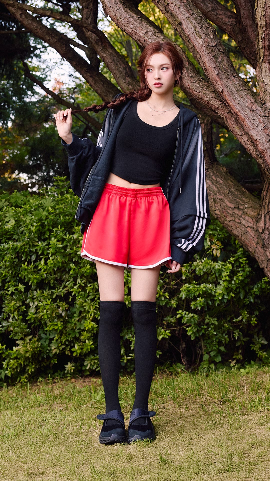 A young woman stands in a grassy area surrounded by trees, wearing a black top, red shorts, and black knee-high socks.