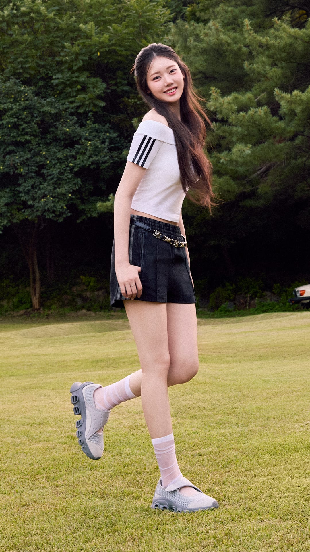 A young woman with long brown hair stands on a grassy field, wearing a white top with stripes and black shorts, with a backdrop of lush green foliage.