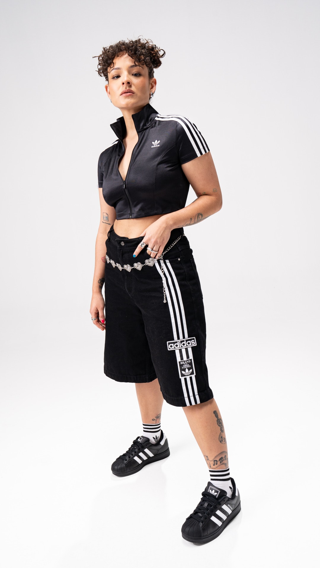 A young person with curly hair wearing a black athletic outfit and Adidas sneakers stands against a plain white background.