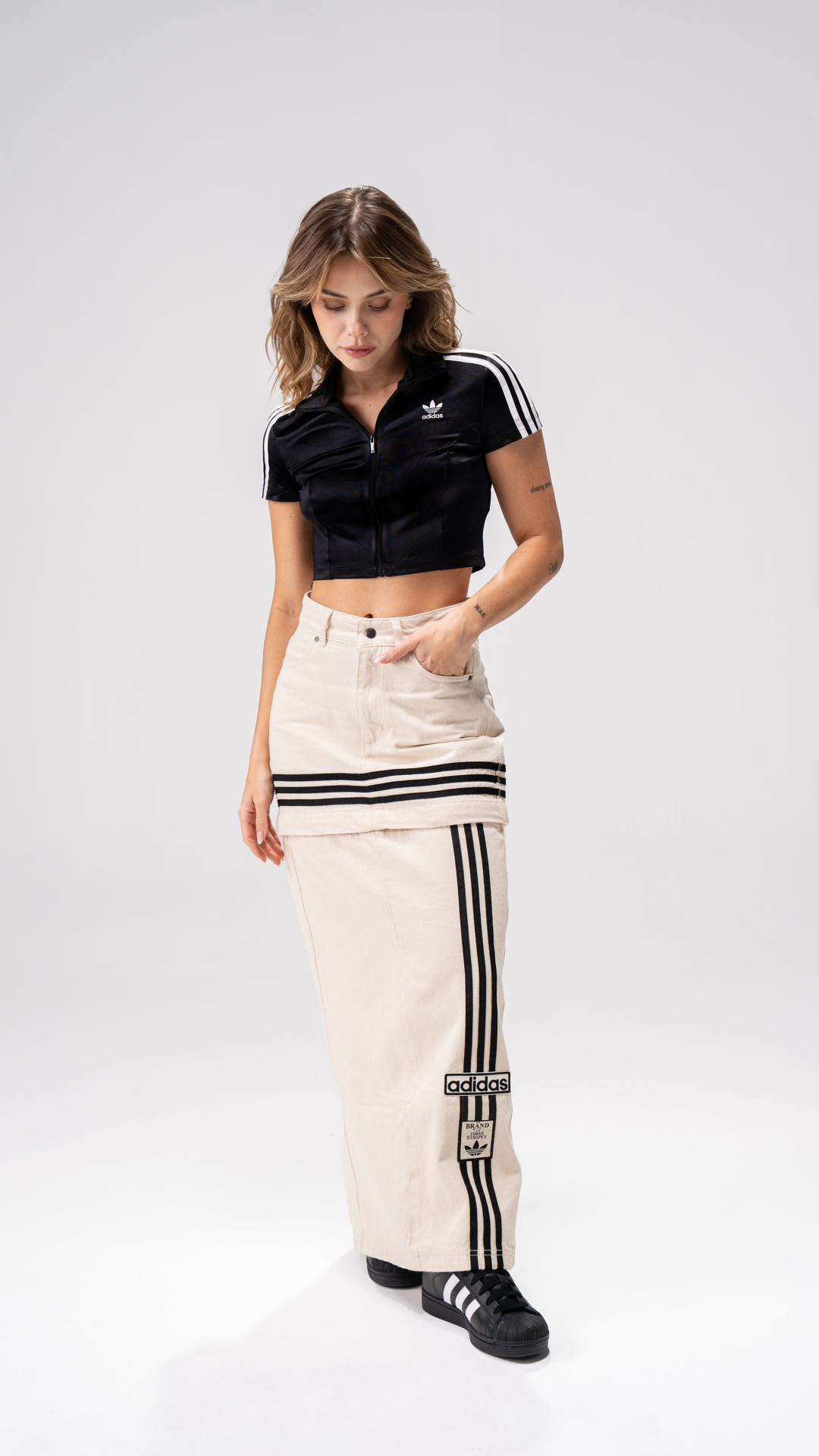 The image shows a young woman wearing a black crop top and white pants with black stripes, standing against a plain white background.