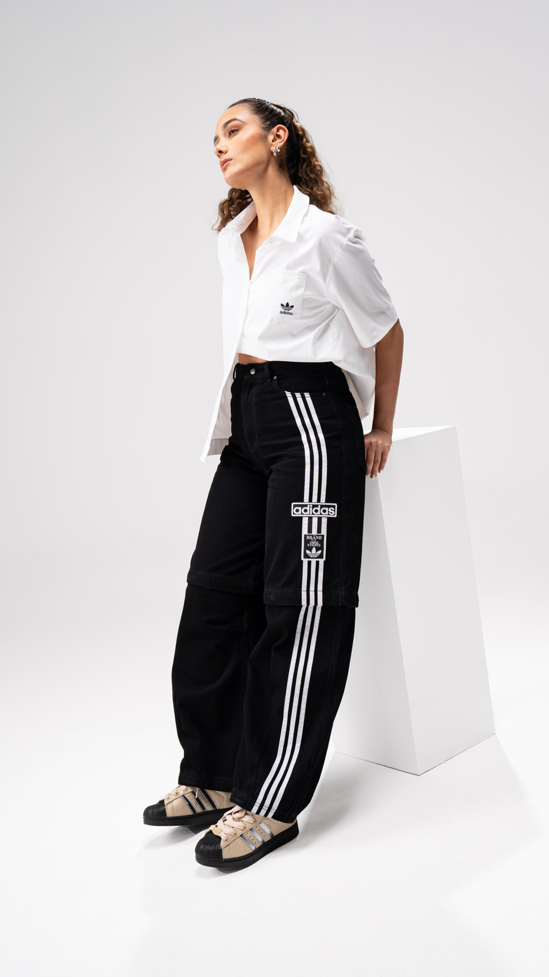 A person wearing a white blouse and black pants with stripes sits on a white platform against a plain background.