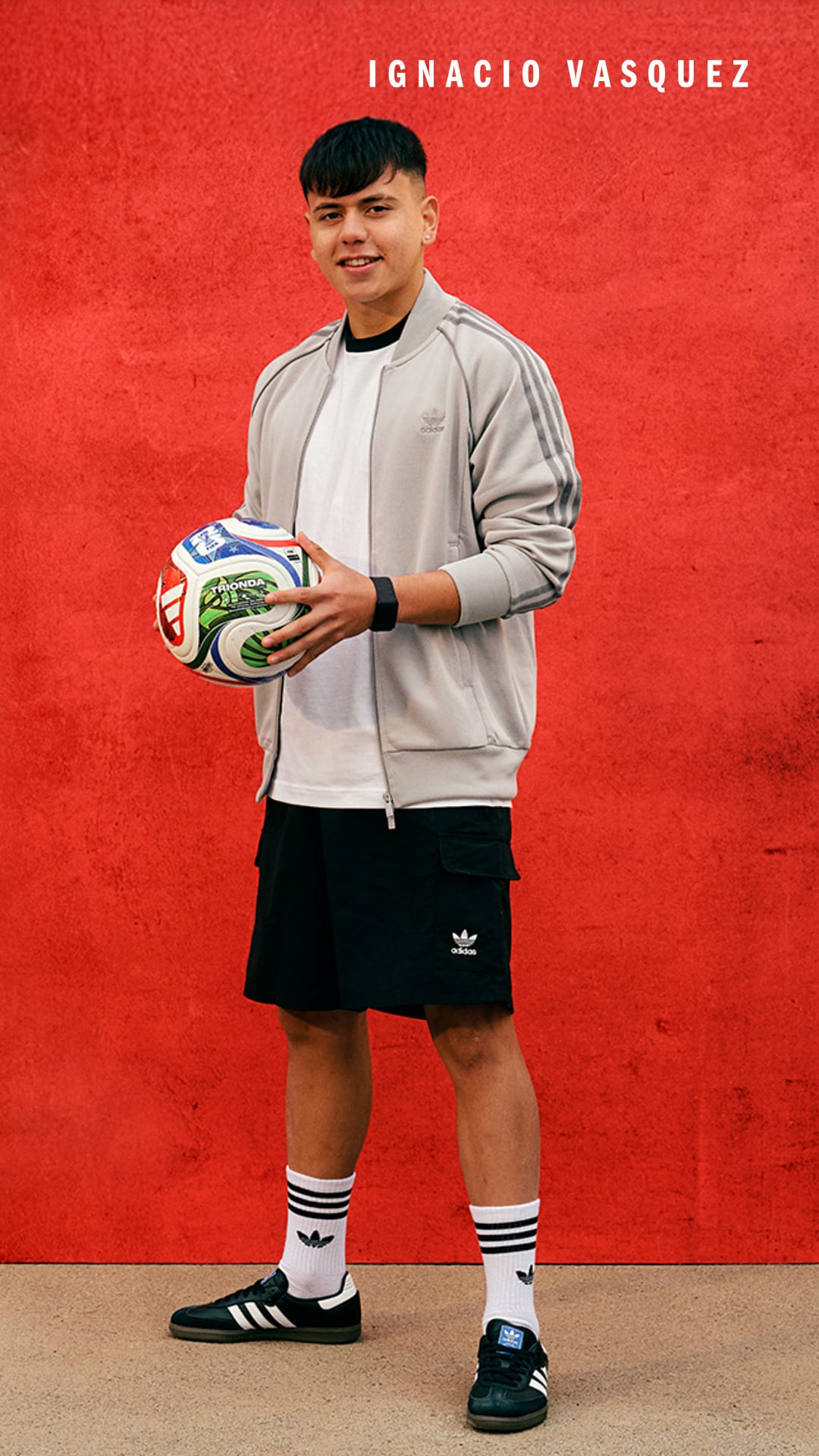 A young man in casual athletic attire stands in front of a vibrant red background, holding a soccer ball.