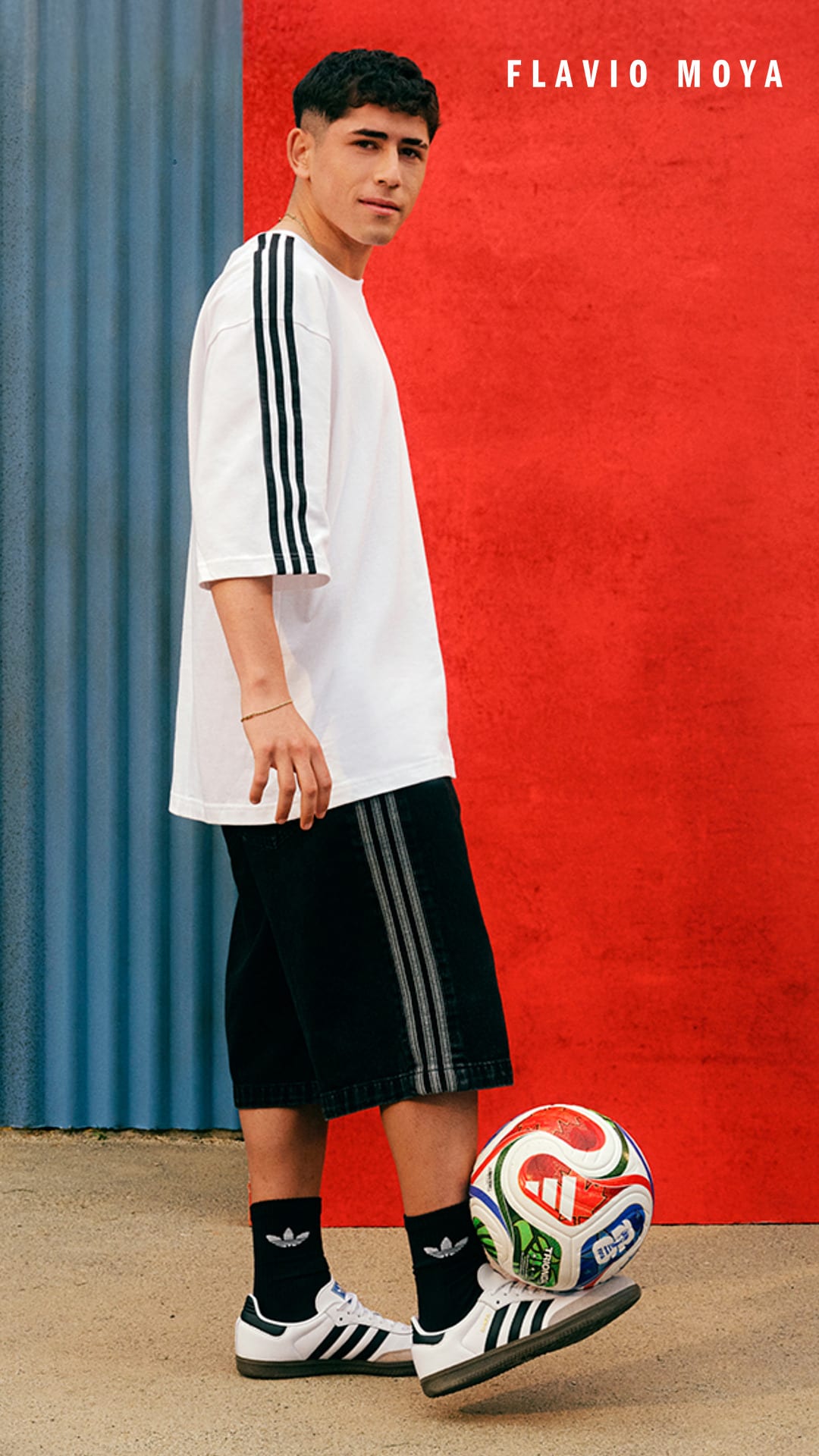 A young man wearing a white t-shirt, black shorts, and Adidas sneakers stands in front of a bright red background with the name "Flavio Moya" printed on it, holding a soccer ball.