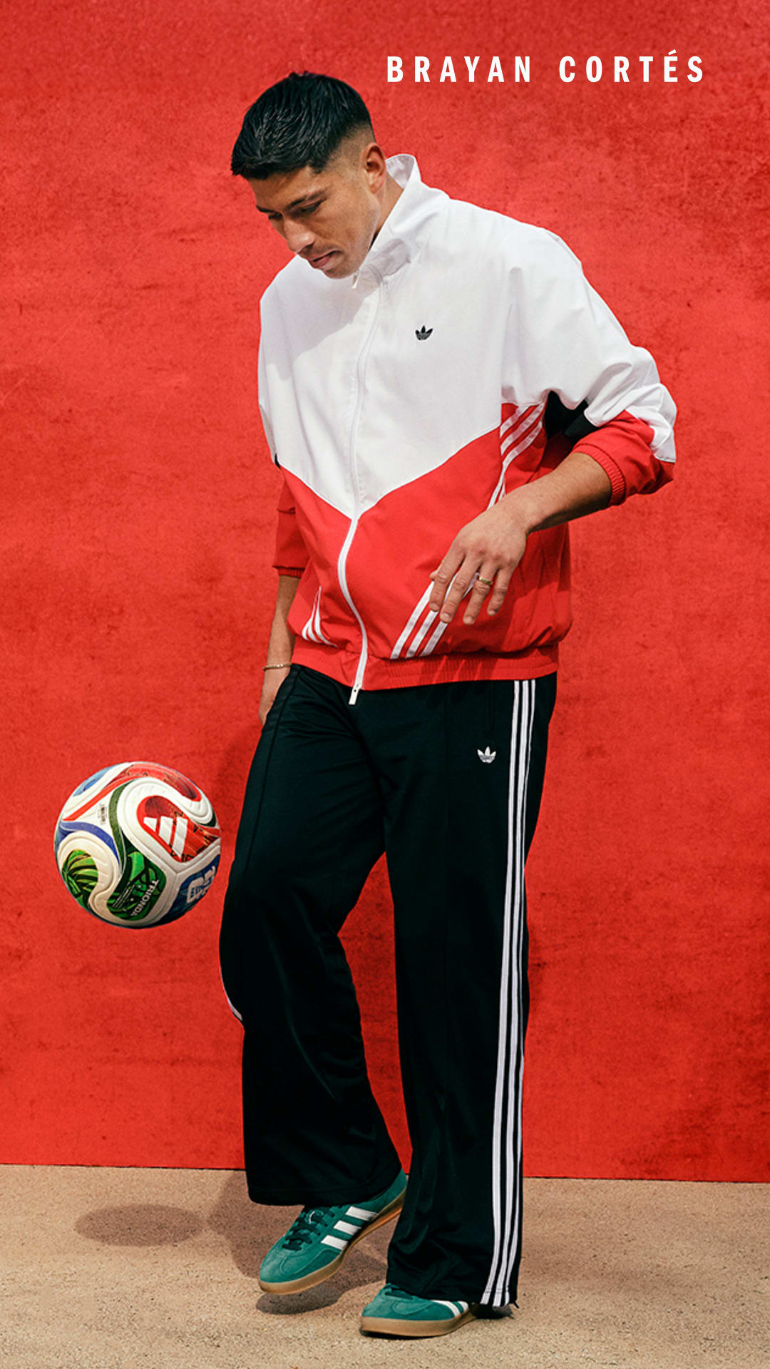 A young man in a white and red athletic jacket stands in front of a bright red background, holding a soccer ball.
