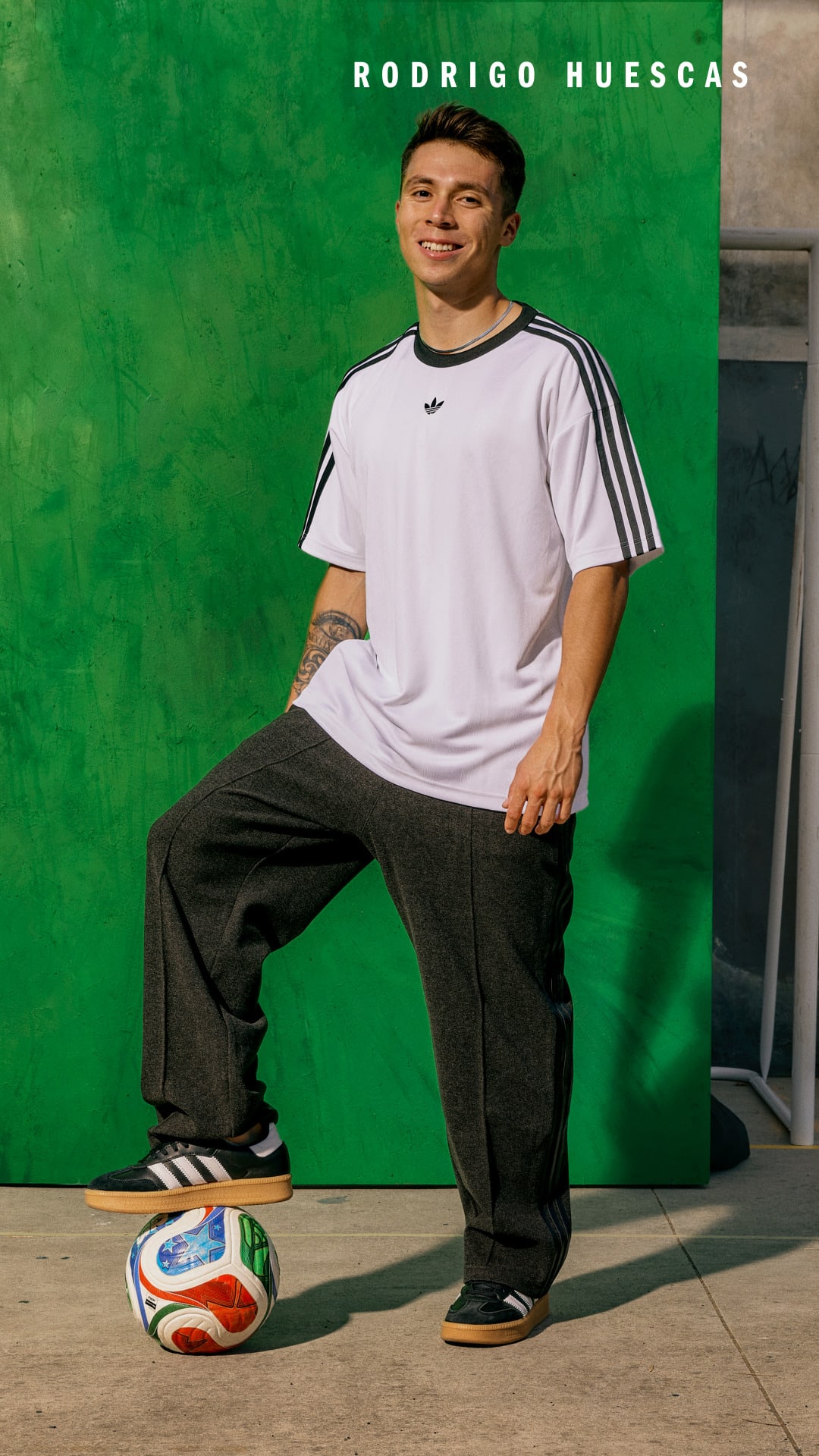 The image shows a young man wearing a white t-shirt and black pants, sitting on the floor next to a soccer ball in front of a green background with the name "RODRIGO HUESCAS" displayed.
