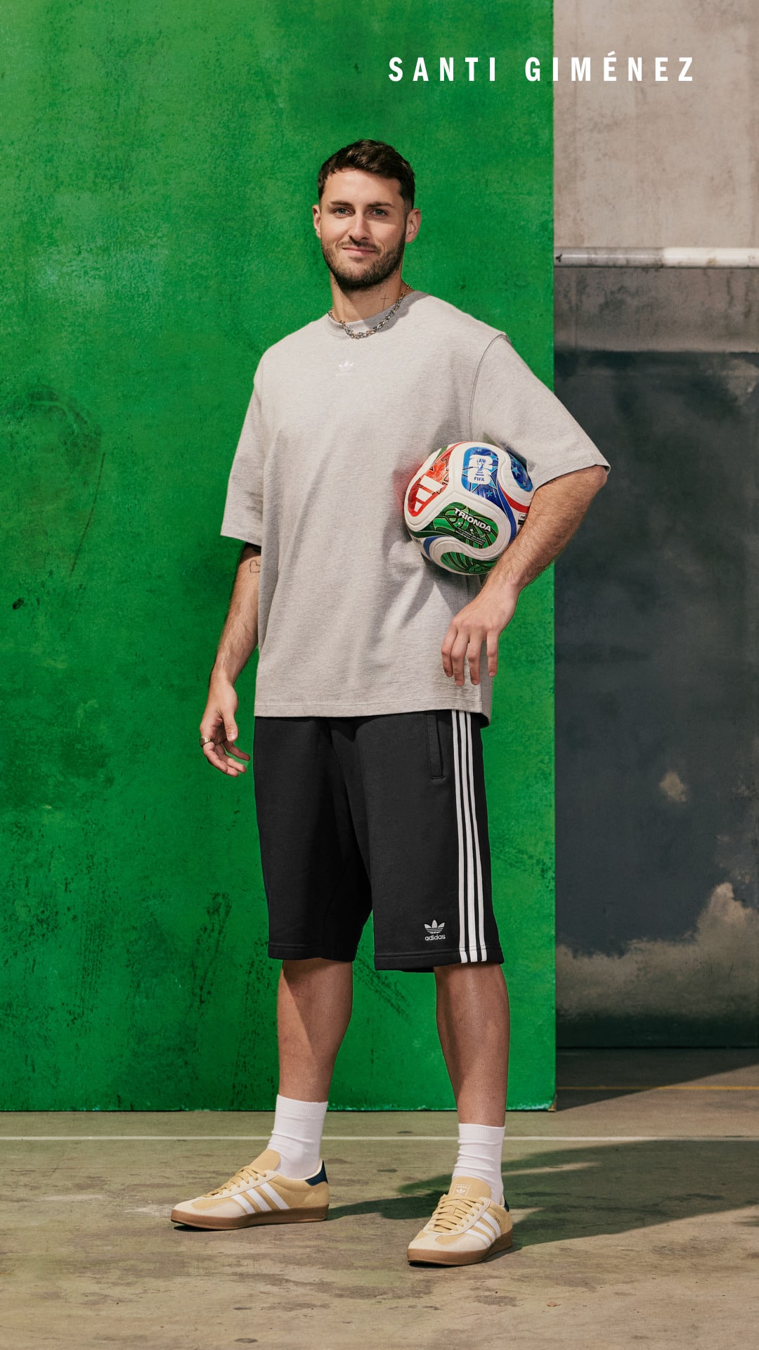 A young man in a white t-shirt and black shorts stands in front of a green background, holding a soccer ball.