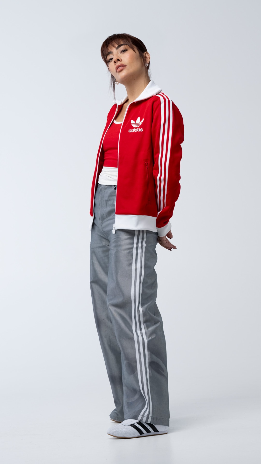 A young person wearing a red Adidas jacket and gray pants with white stripes, standing against a plain white background.