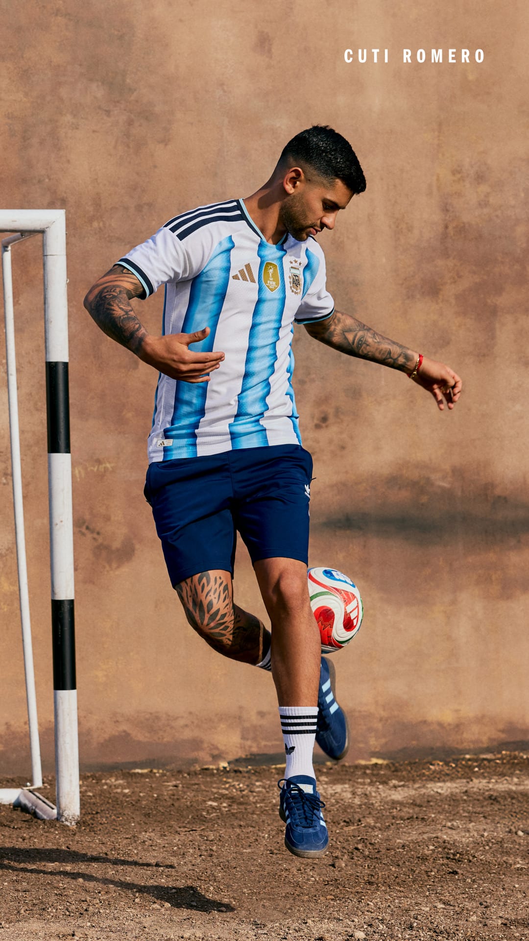 A soccer player in an Argentina jersey is dribbling a ball on a dirt field with a plain background.