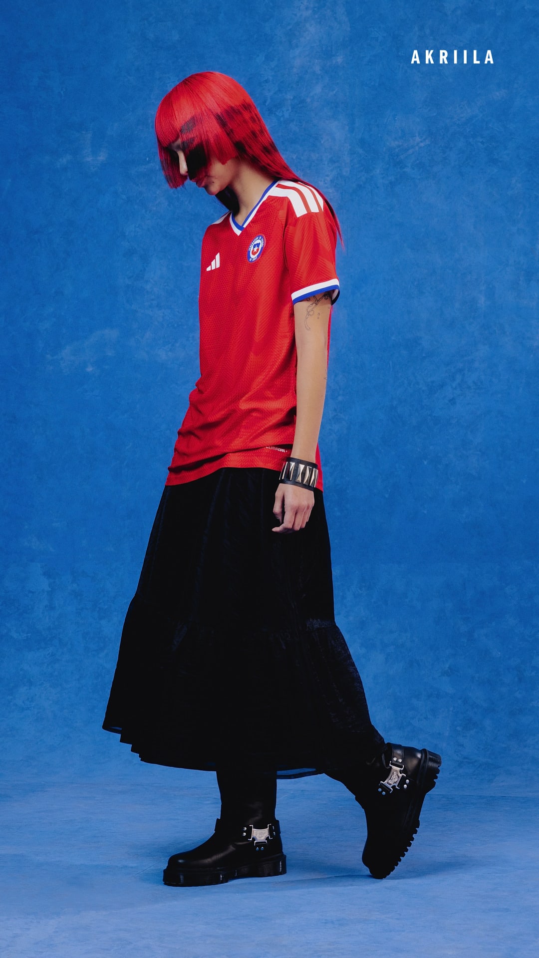 A person wearing a red jersey and black skirt stands against a vibrant blue background.