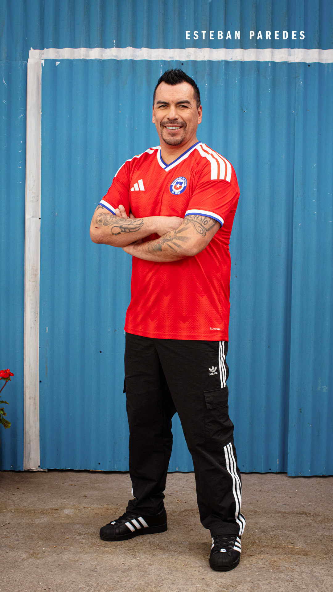 A smiling man wearing a red soccer jersey and black pants stands in front of a blue corrugated metal wall.