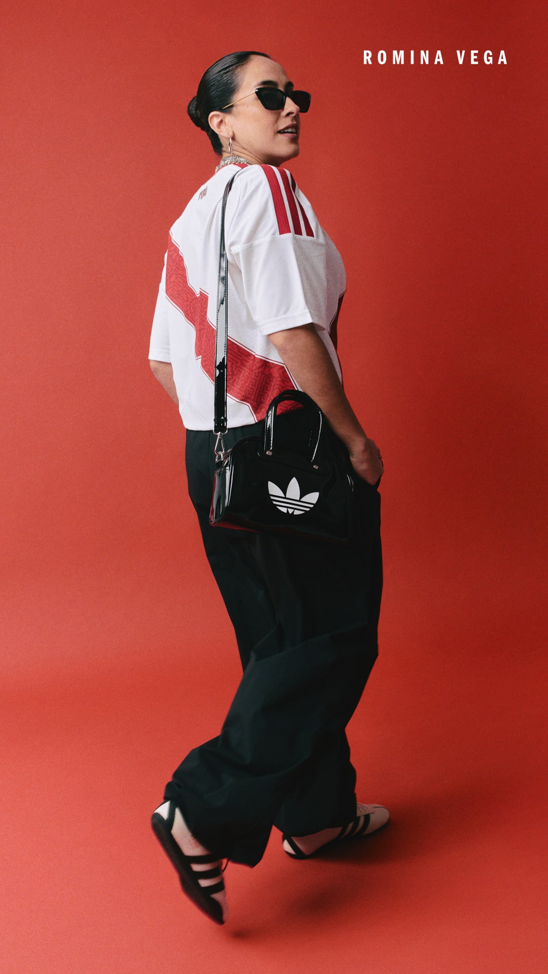 A person wearing a white top with red stripes, black pants, and sunglasses stands against a vibrant red background.