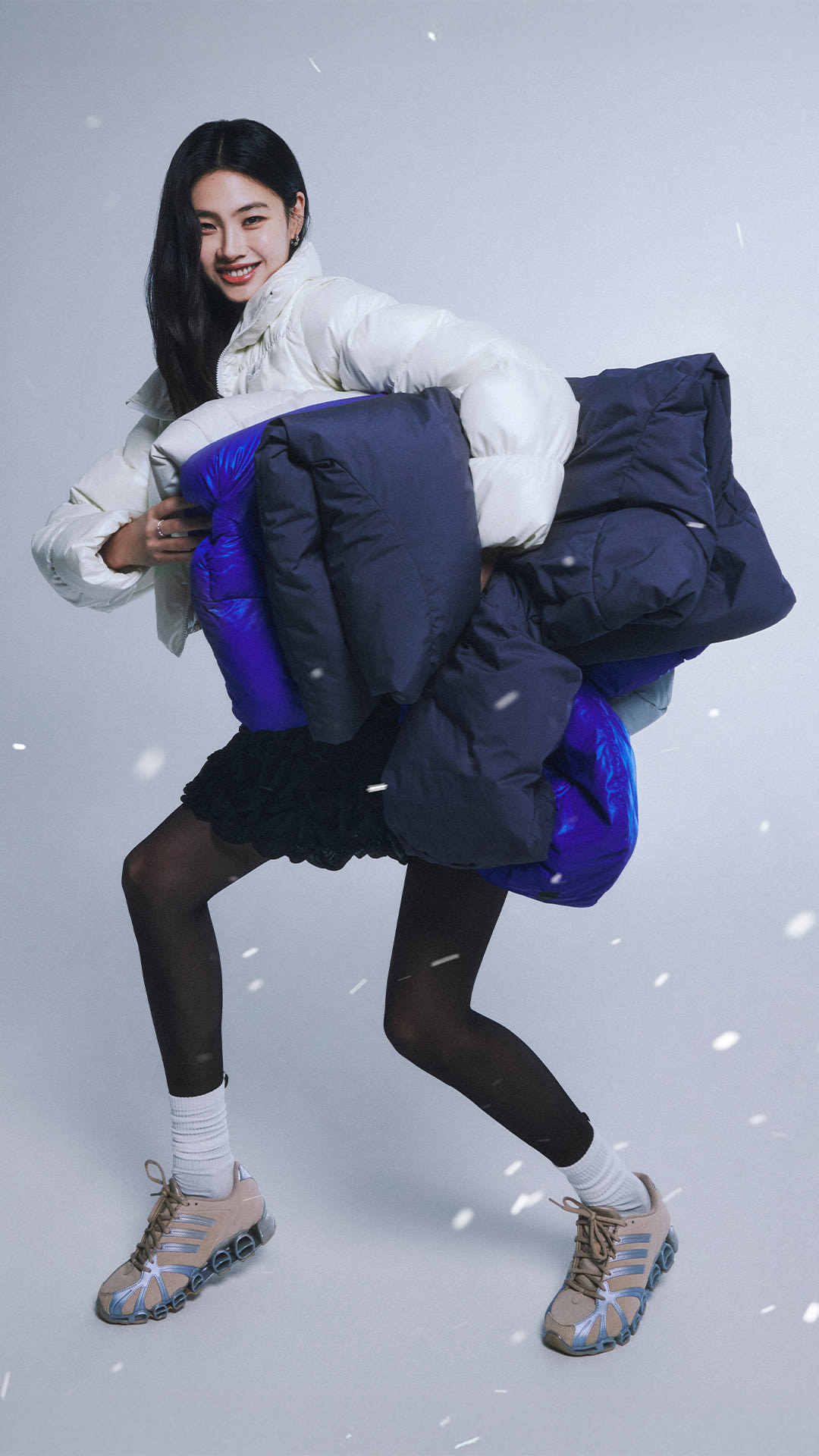 A young woman in a white fur-trimmed jacket and navy blue puffer jacket is crouching in the snow, surrounded by a snowy background.