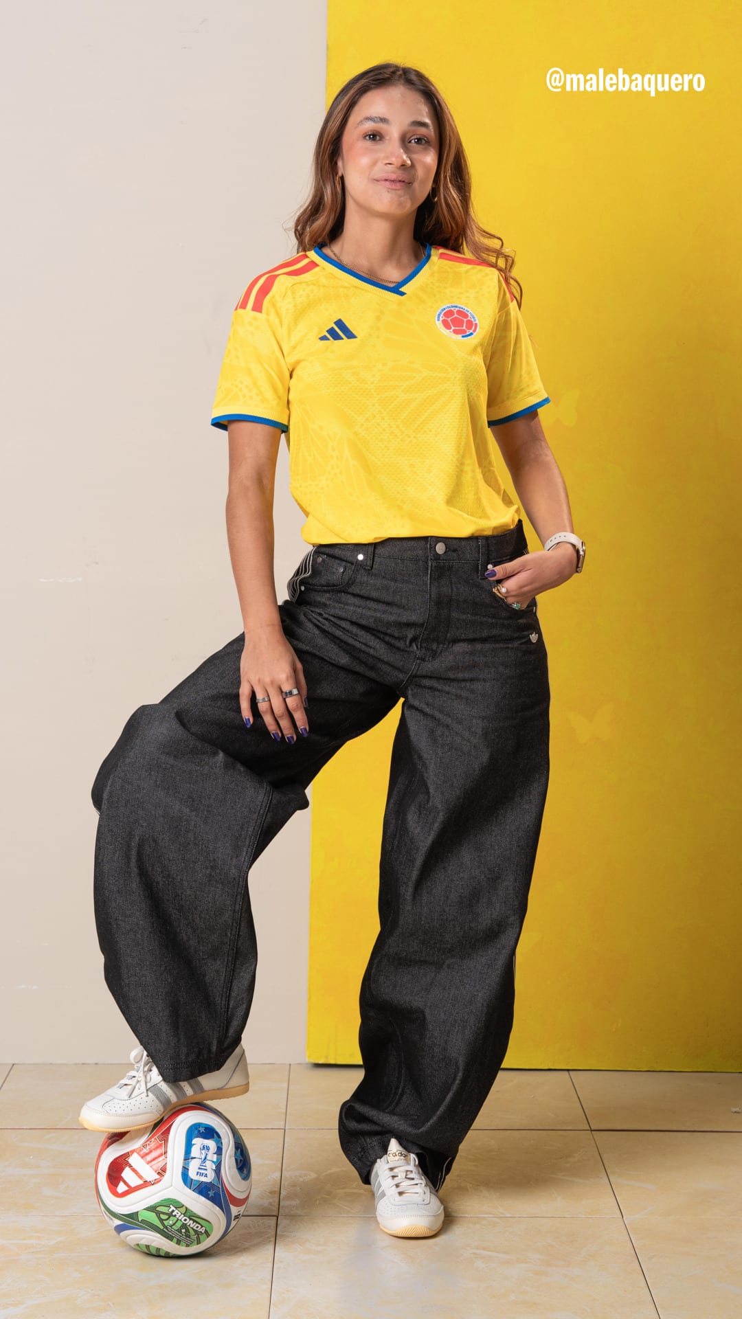 A young woman wearing a yellow soccer jersey and black pants stands in front of a yellow background, posing with a soccer ball at her feet.