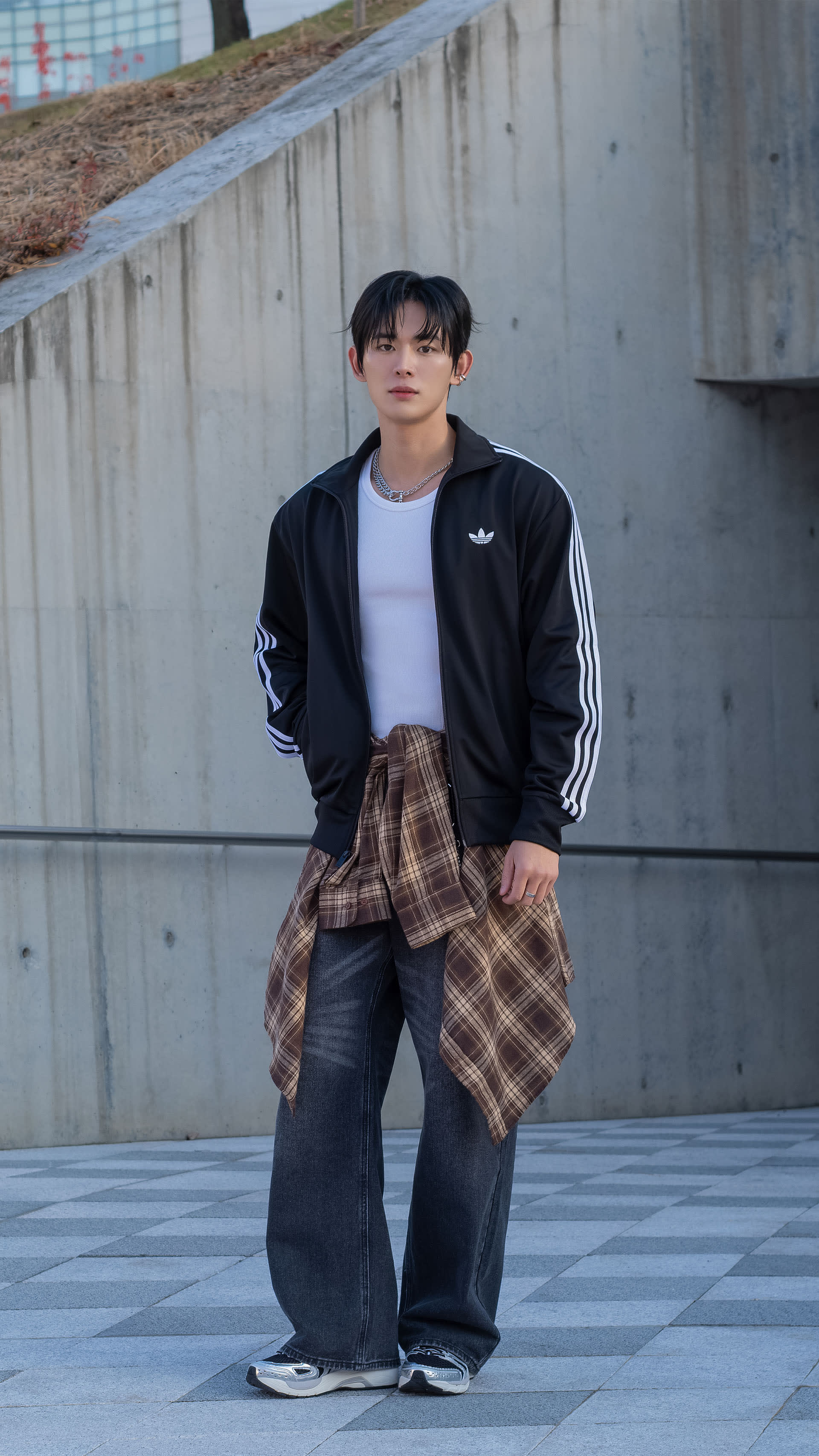 A young person wearing a black jacket and plaid pants stands in front of a concrete wall in an urban setting.