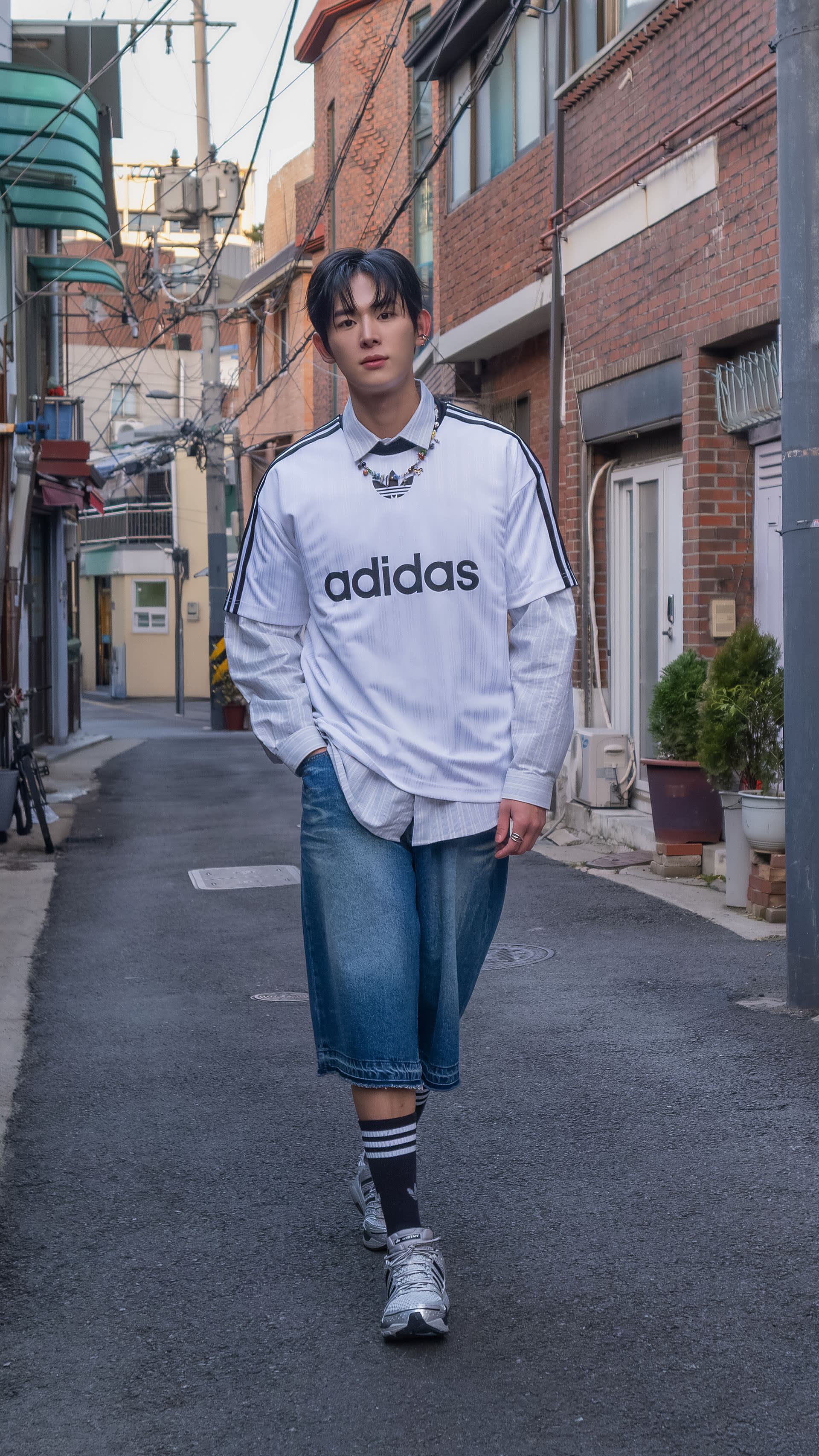 A person wearing an Adidas sweatshirt and jeans is standing on a street with brick buildings and storefronts in the background.