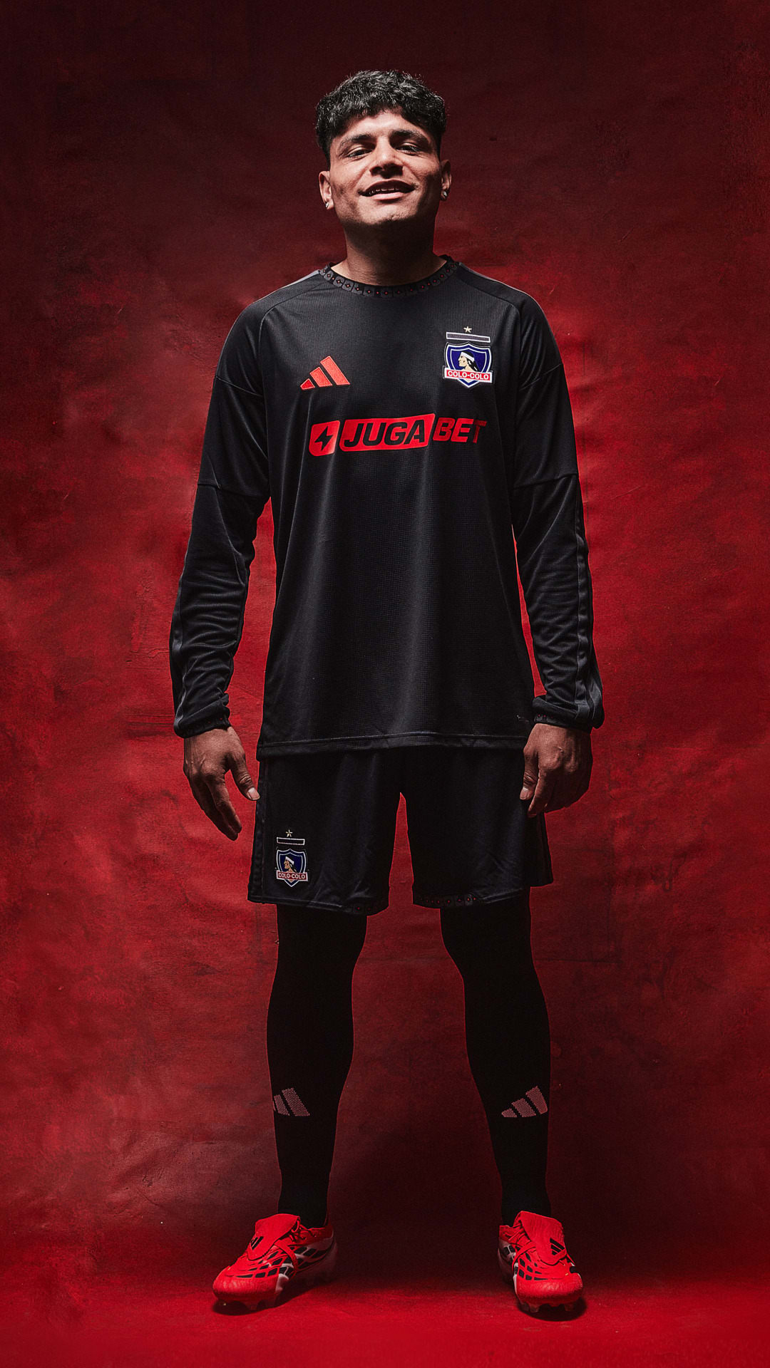 A young man wearing a black sports jersey and shorts stands in front of a vibrant red background, his expression conveying a sense of confidence and determination.