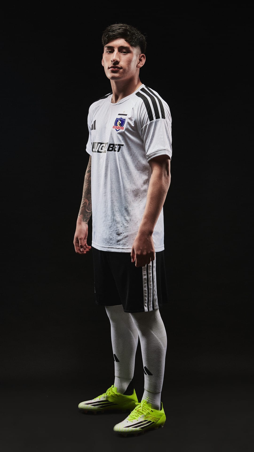 A young man wearing a white soccer jersey and black shorts stands against a dark background, his feet adorned with bright green cleats.