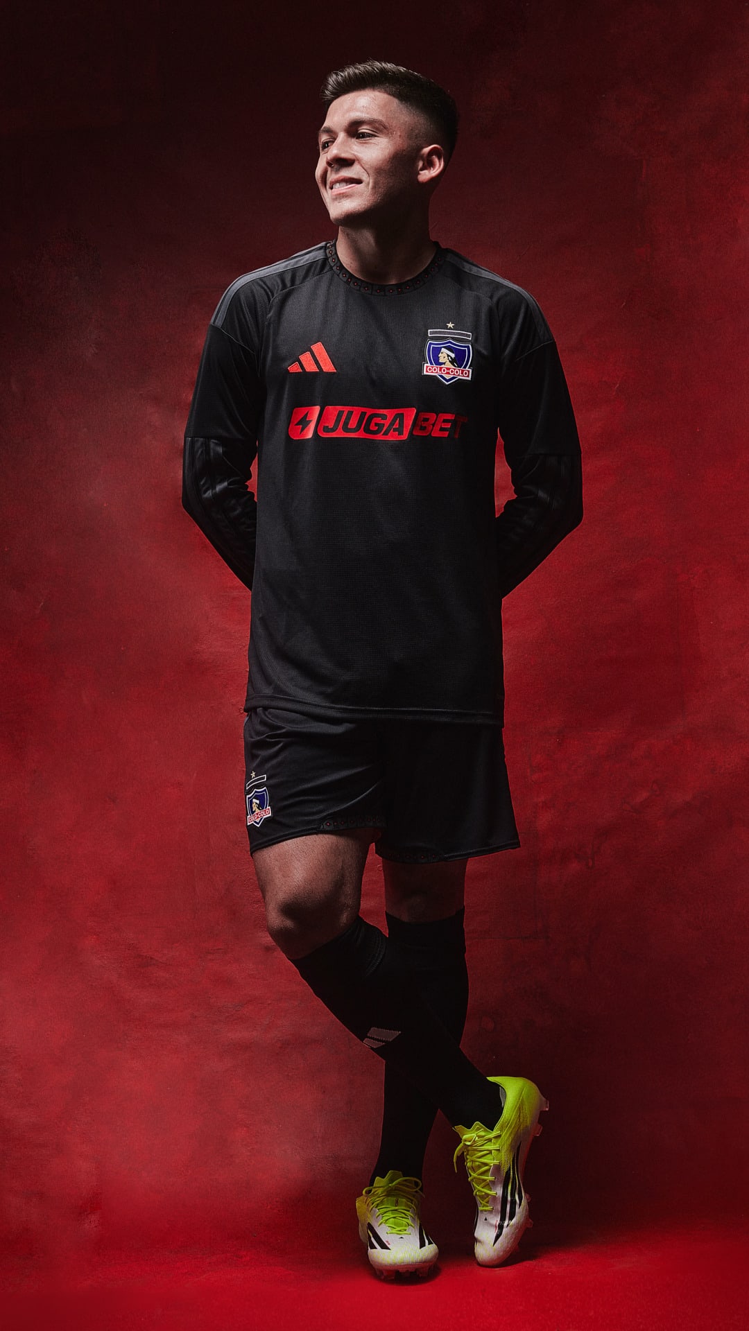 A young man wearing a black sports jersey with the team logo "Johor" stands against a deep red background, his bright yellow cleats adding a vibrant contrast to the scene.