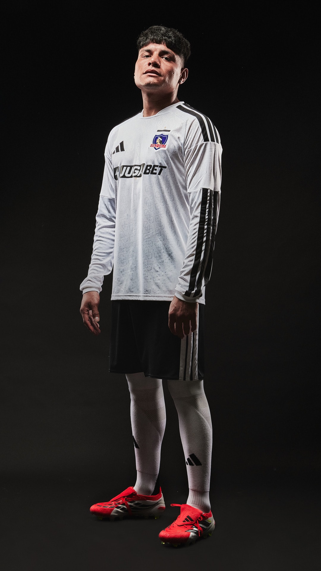 A young man wearing a white and black soccer jersey stands in a dark background, with his hands on his hips and a serious expression on his face.