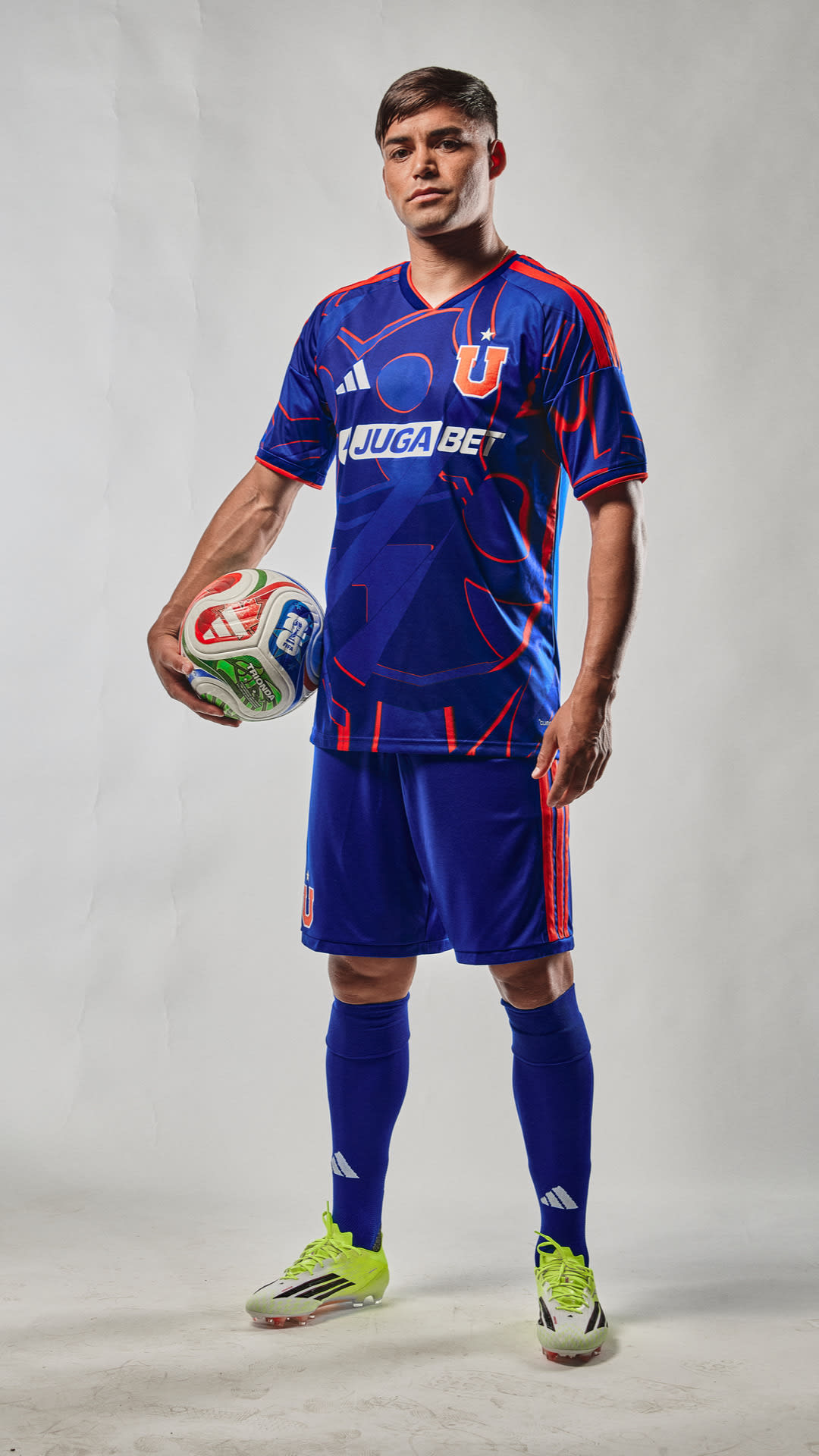 A man wearing a blue and red soccer jersey and shorts, holding a soccer ball, stands against a plain background.