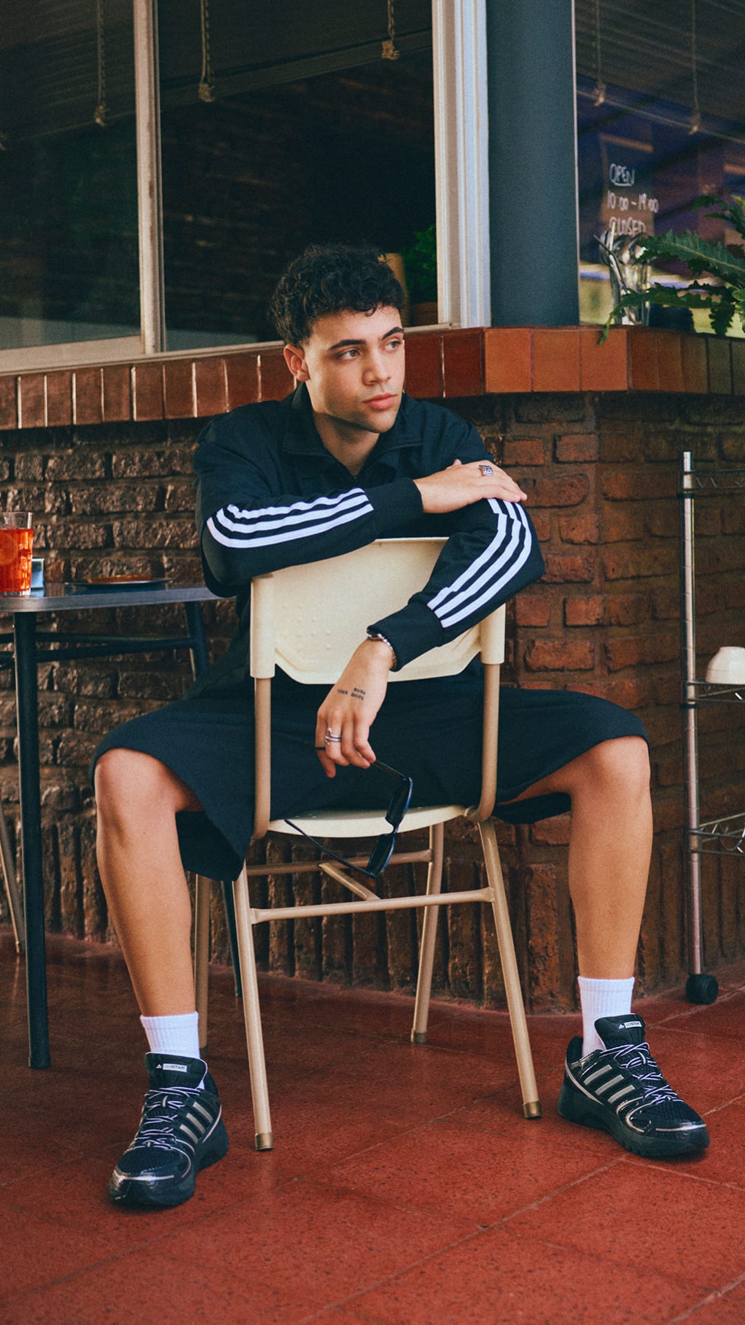 A young man in casual attire sits on a chair in what appears to be a restaurant or cafe setting, with a brick wall and other furnishings visible in the background.