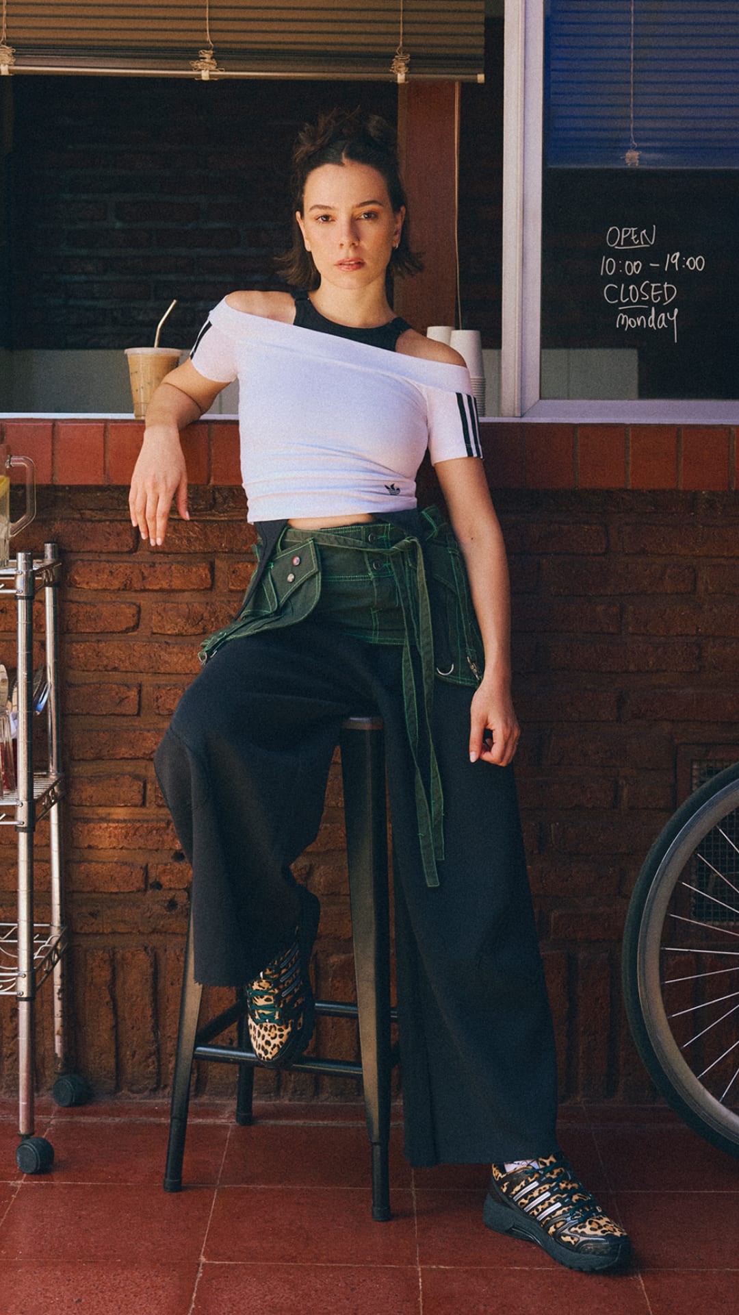 A young woman with dark hair sits on a stool in front of a brick wall, wearing a white top and dark pants, with a bicycle visible in the background.
