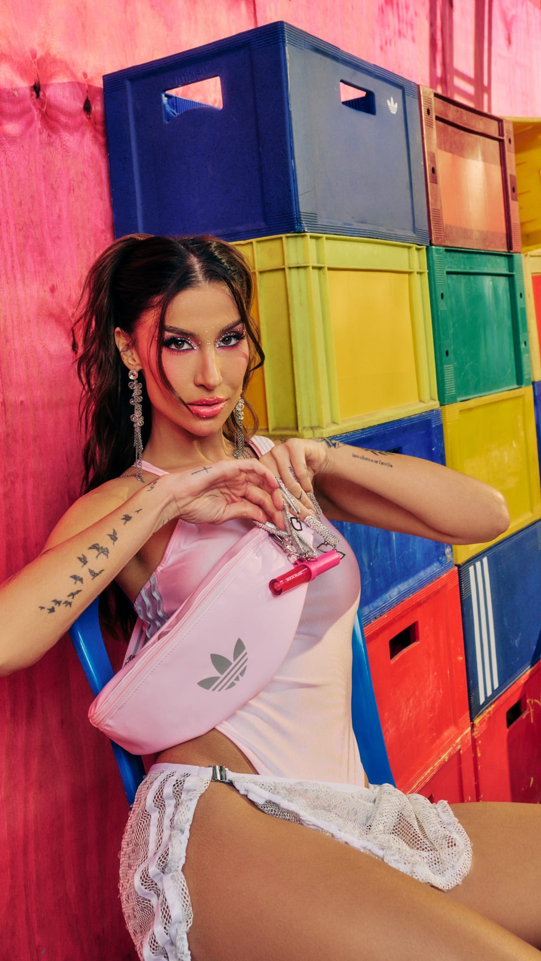 A young woman with curly hair and tattoos poses in front of a colorful backdrop of stacked crates.