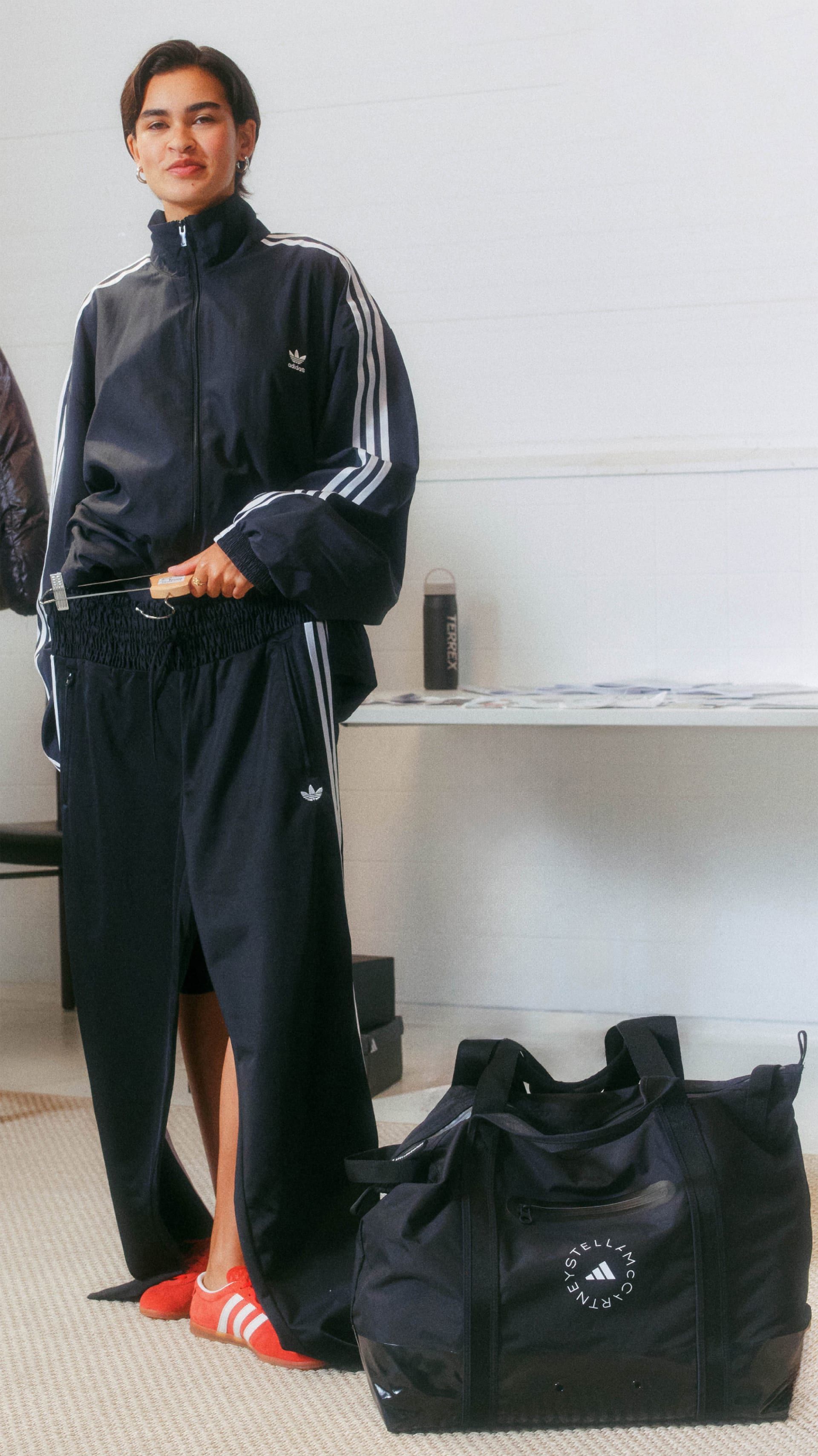 A person wearing an Adidas tracksuit stands in a room with a white wall and a bag on the floor.