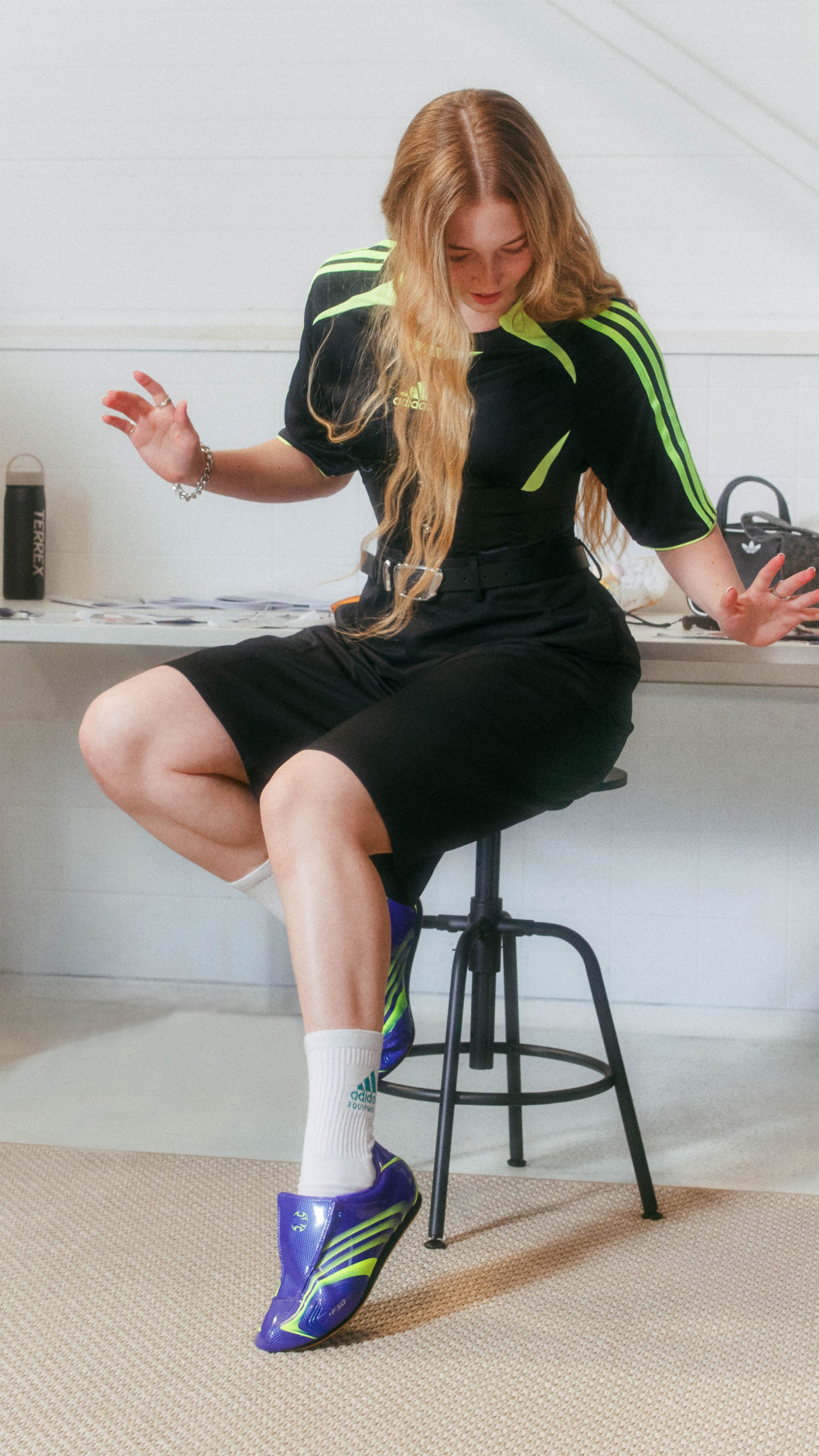 A young woman with long, wavy blonde hair is sitting on a stool, wearing a black and green dress and purple sneakers, in what appears to be a home or office setting.