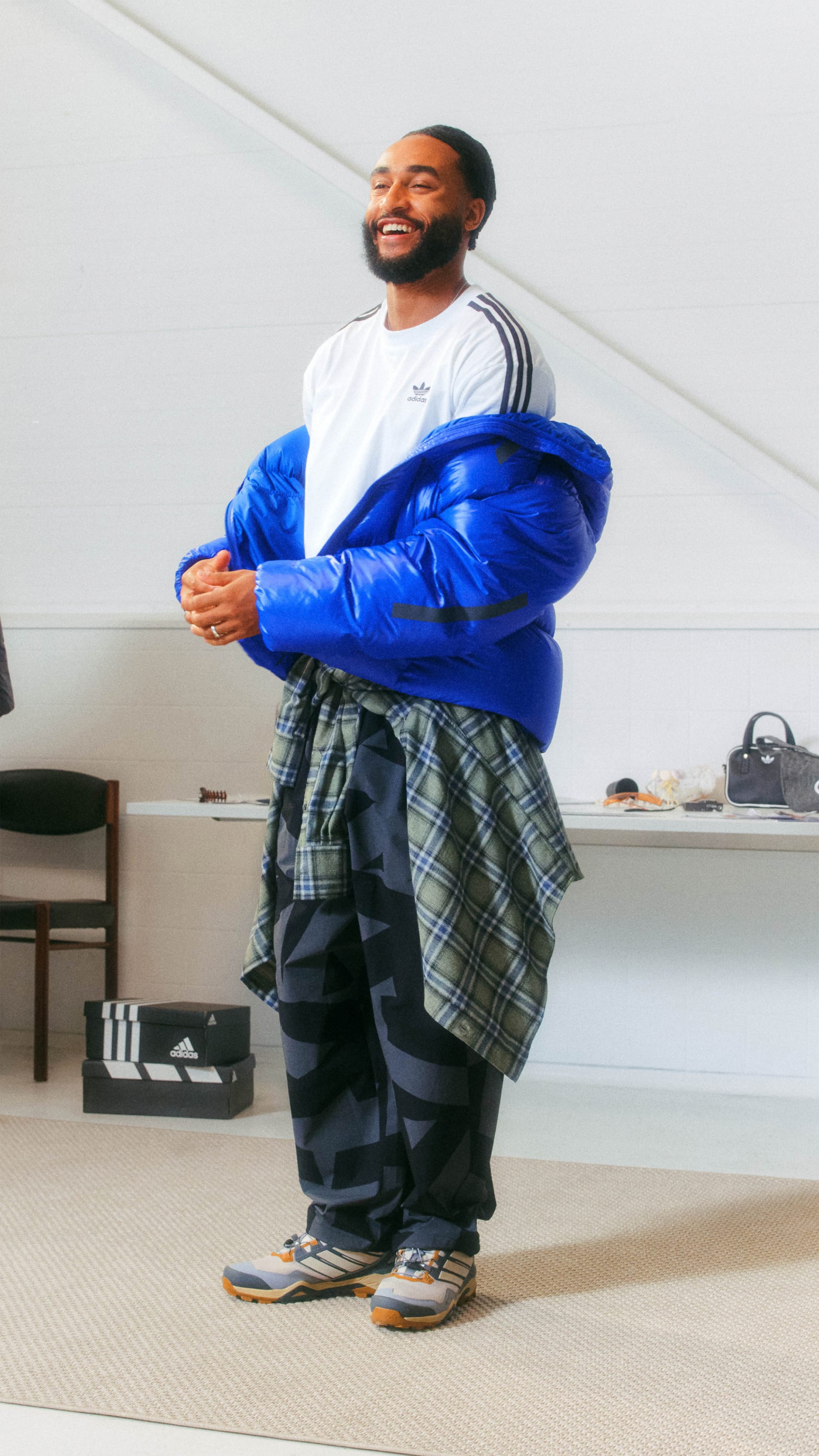 A smiling man wearing a white shirt, blue jacket, and checkered pants stands in a room with shelves and furniture in the background.