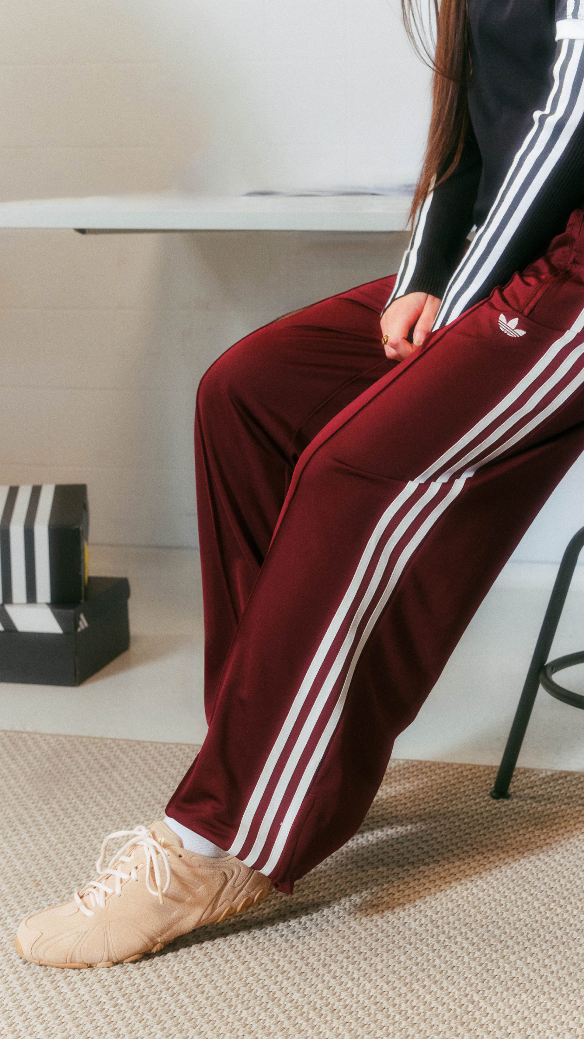 A person wearing a red and white striped sweatpants and white sneakers is sitting on a chair in a room with a white wall and a black object in the background.