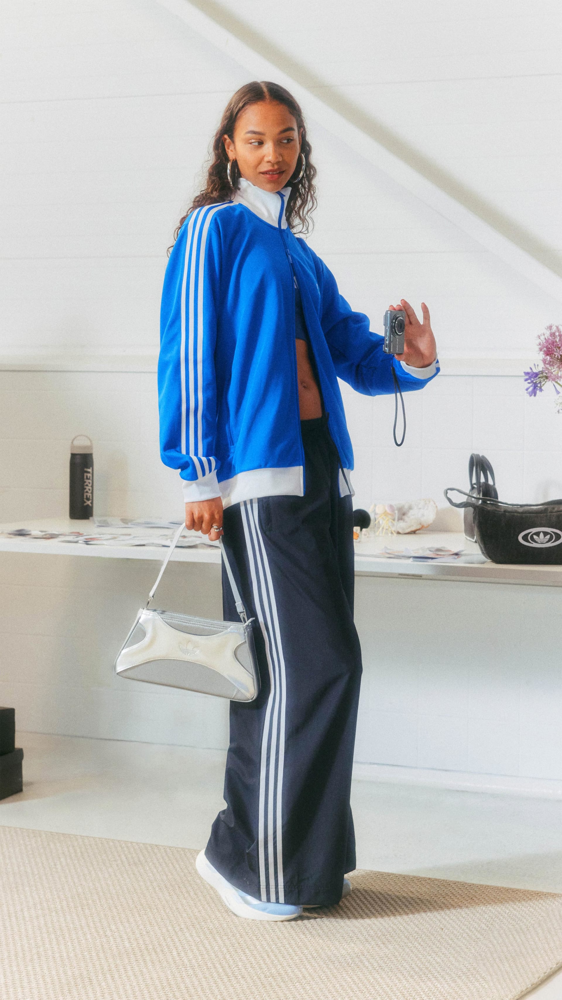 A young woman wearing a blue jacket and navy blue pants stands in a kitchen, with various kitchen appliances visible in the background.