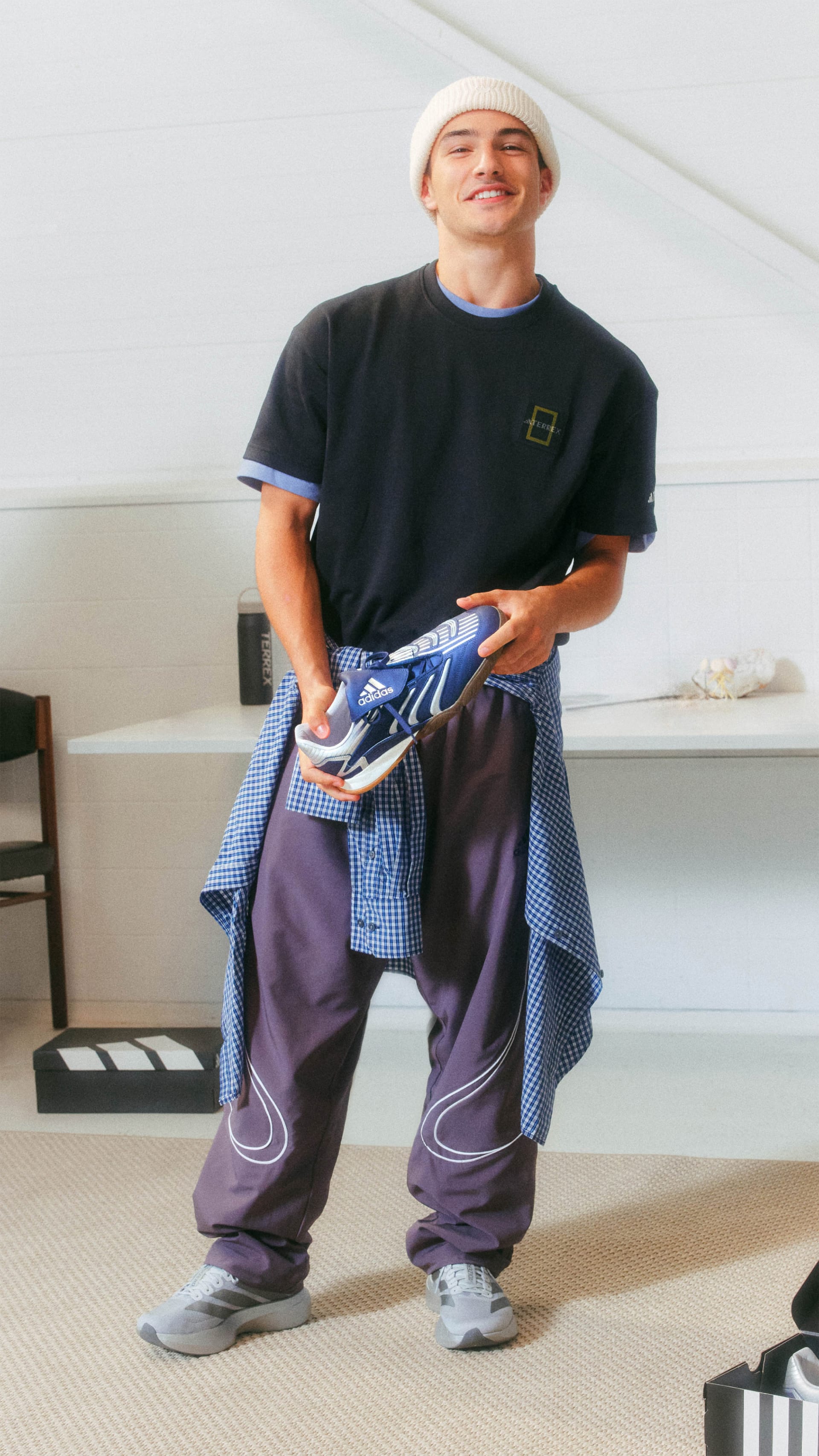 A smiling person wearing a black t-shirt, blue and white checkered shirt, and purple pants, holding a video game controller in a room with various objects in the background.