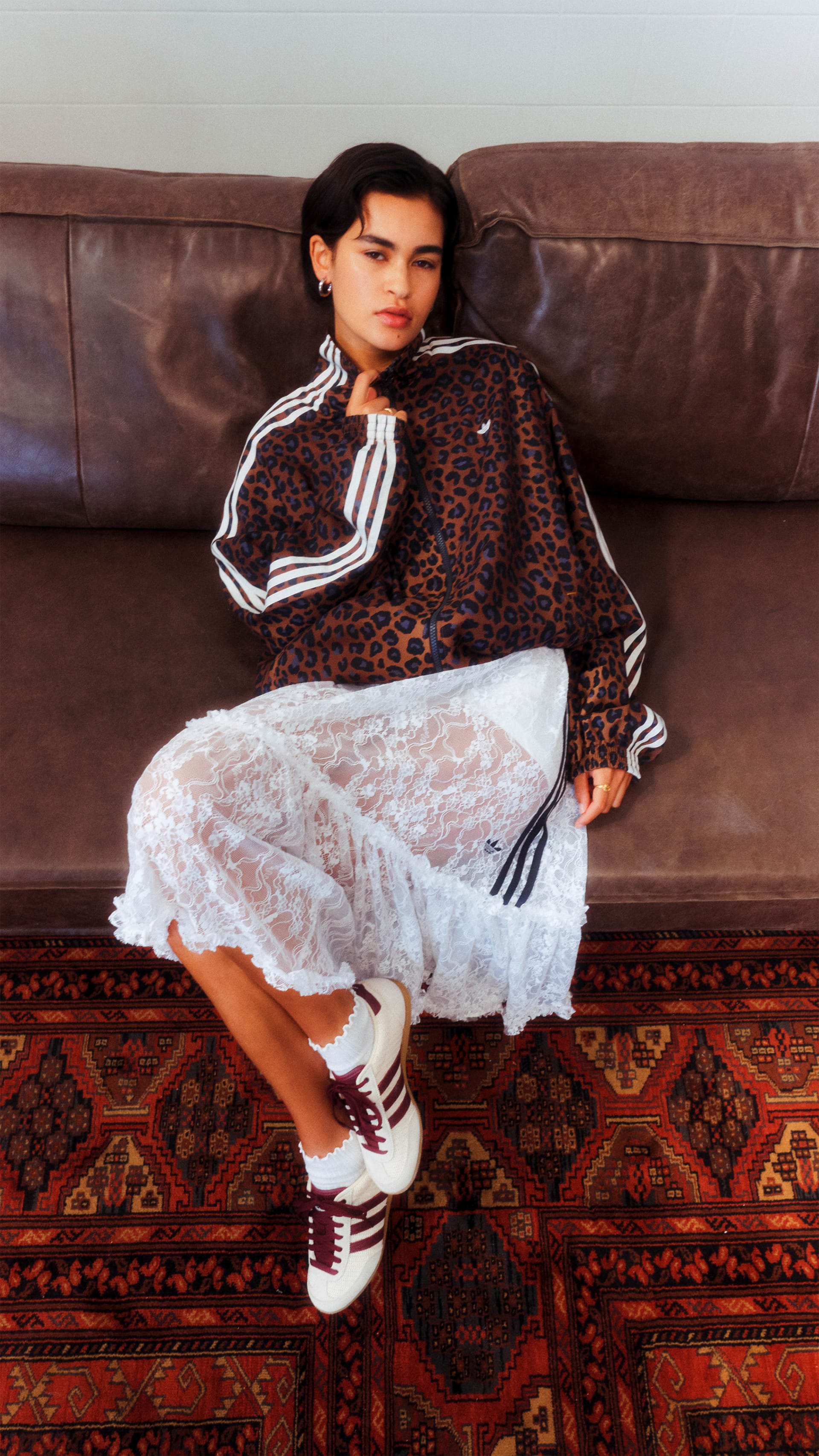 A young woman wearing a leopard print jacket and white lace dress sits on a patterned rug, her legs crossed and her expression pensive.