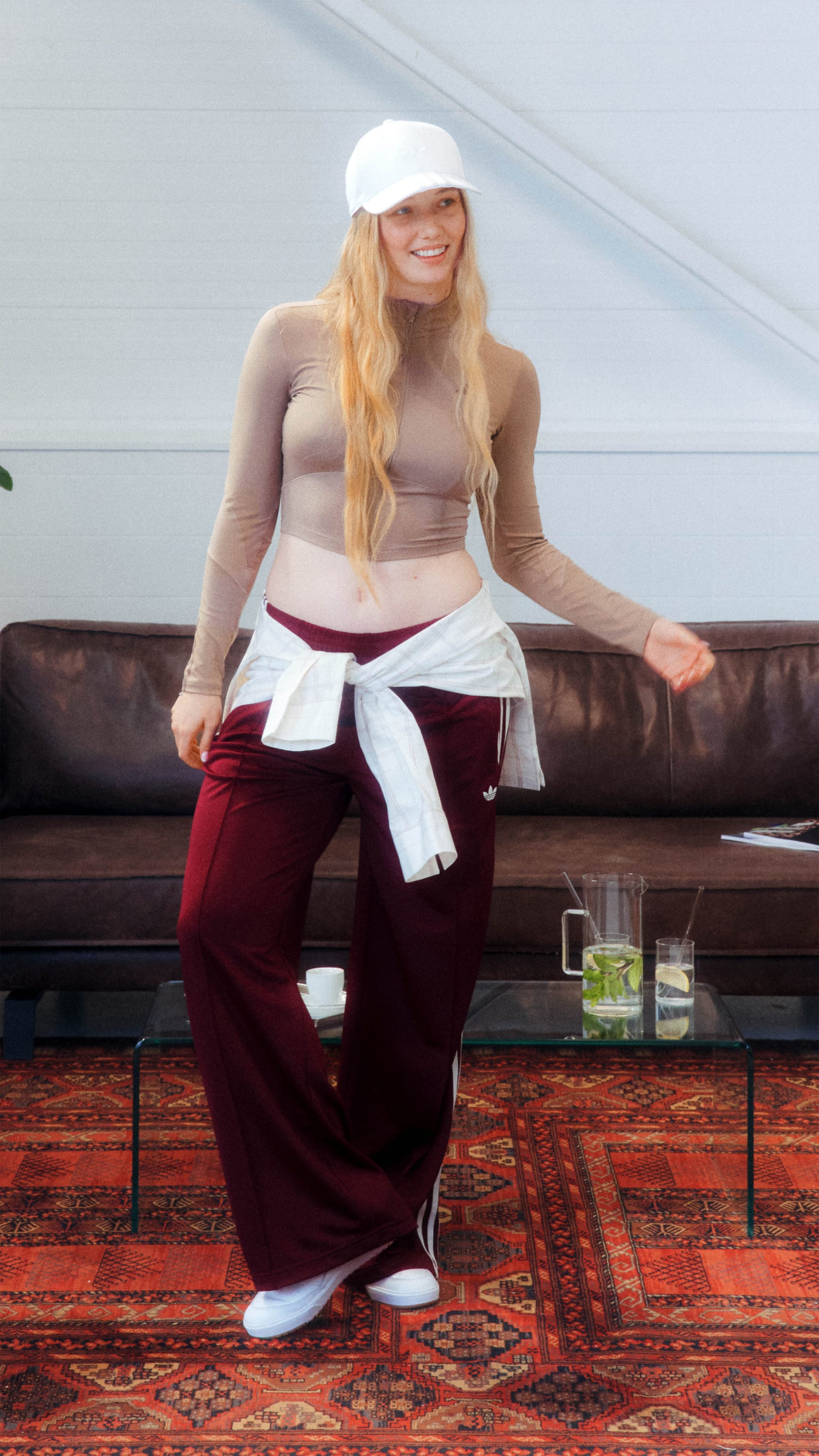 A young woman with long blonde hair sits on a leather couch, wearing a white top, red pants, and a white headband. The background includes a patterned rug and a glass table with plants.