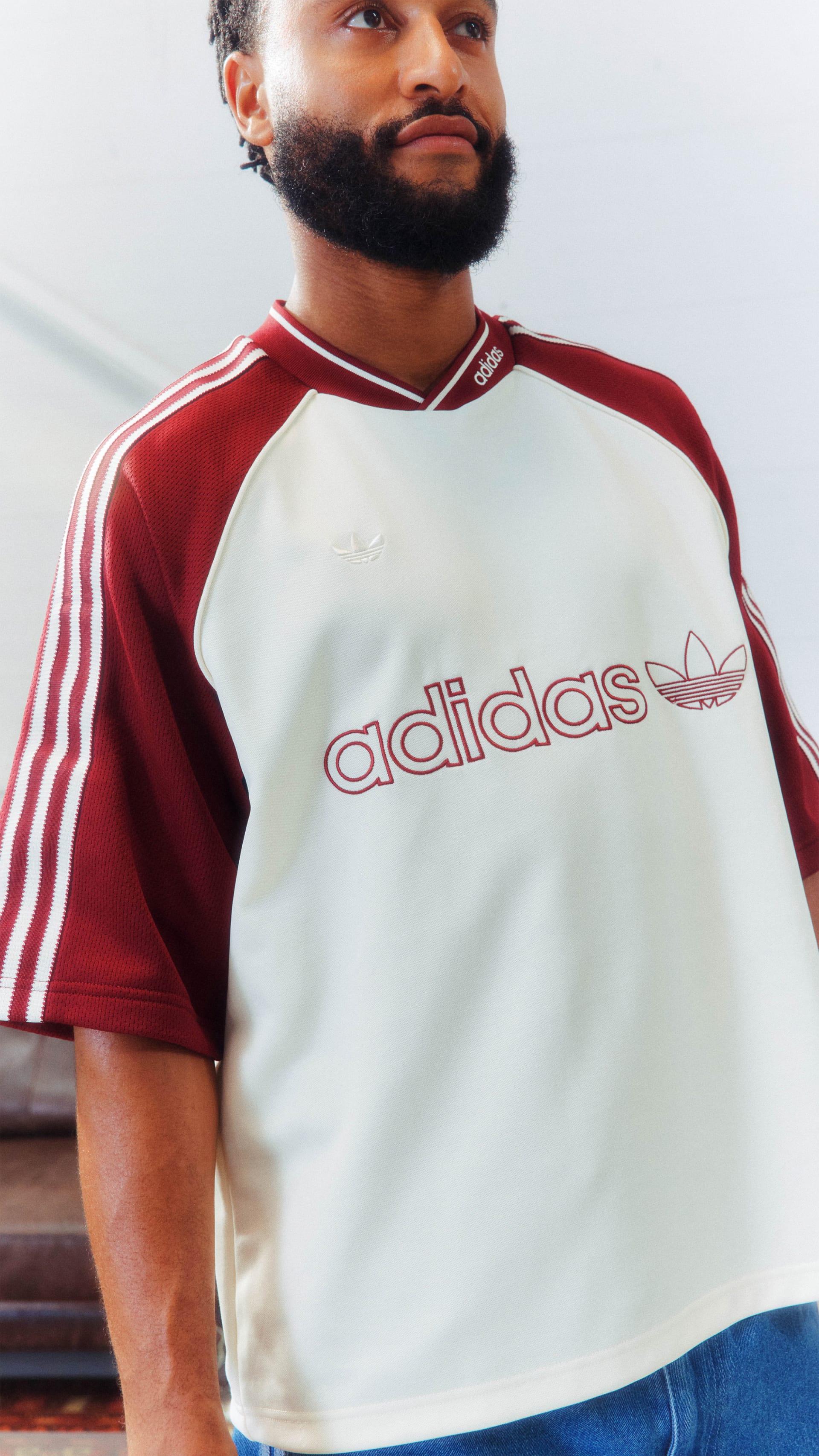 A man with a beard wearing a red and white Adidas jersey stands in front of a plain background.