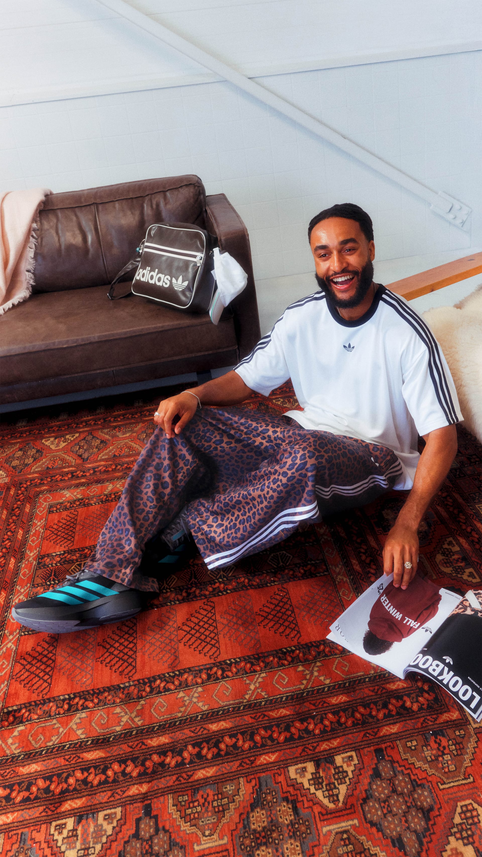 A smiling man sits on a colorful patterned rug in a room with a brown leather sofa and Adidas-branded items in the background.