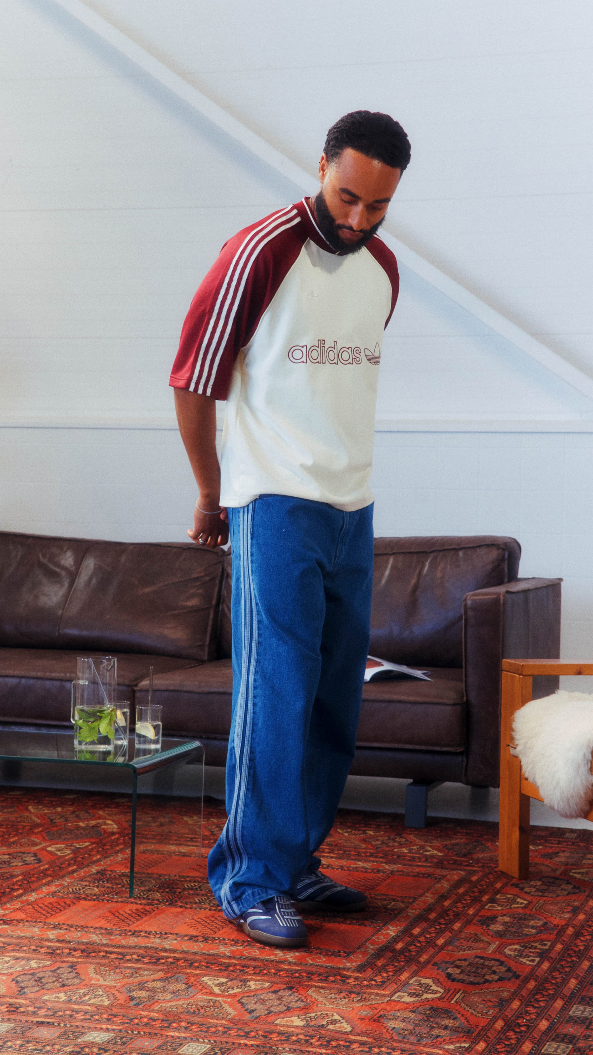 A person wearing a red and white Adidas shirt and blue pants is standing in a room with a leather couch and a rug on the floor.
