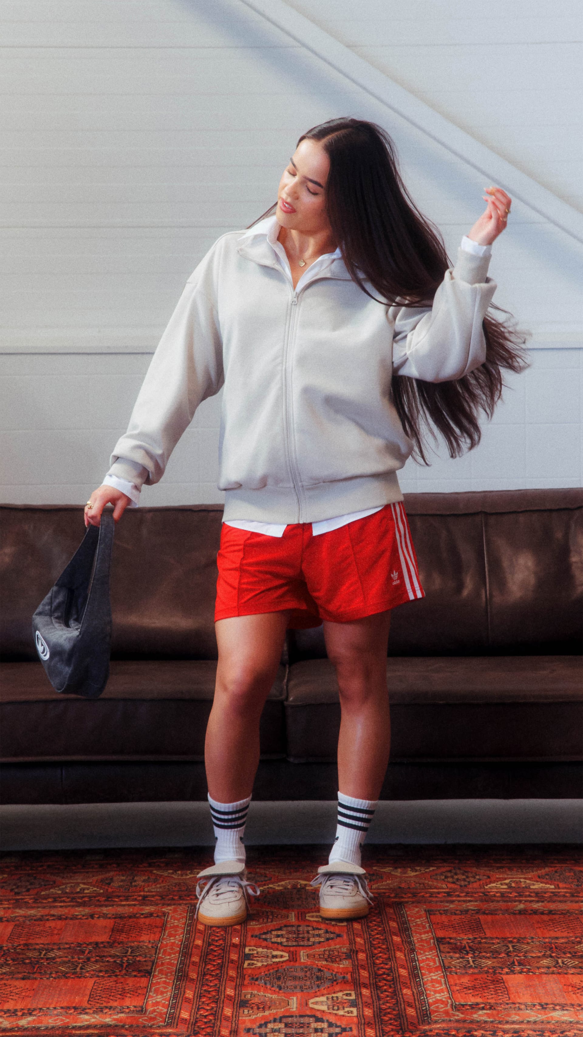 A young woman with long dark hair is standing on a patterned rug, wearing a white sweater and red shorts, her hair blowing in the wind.