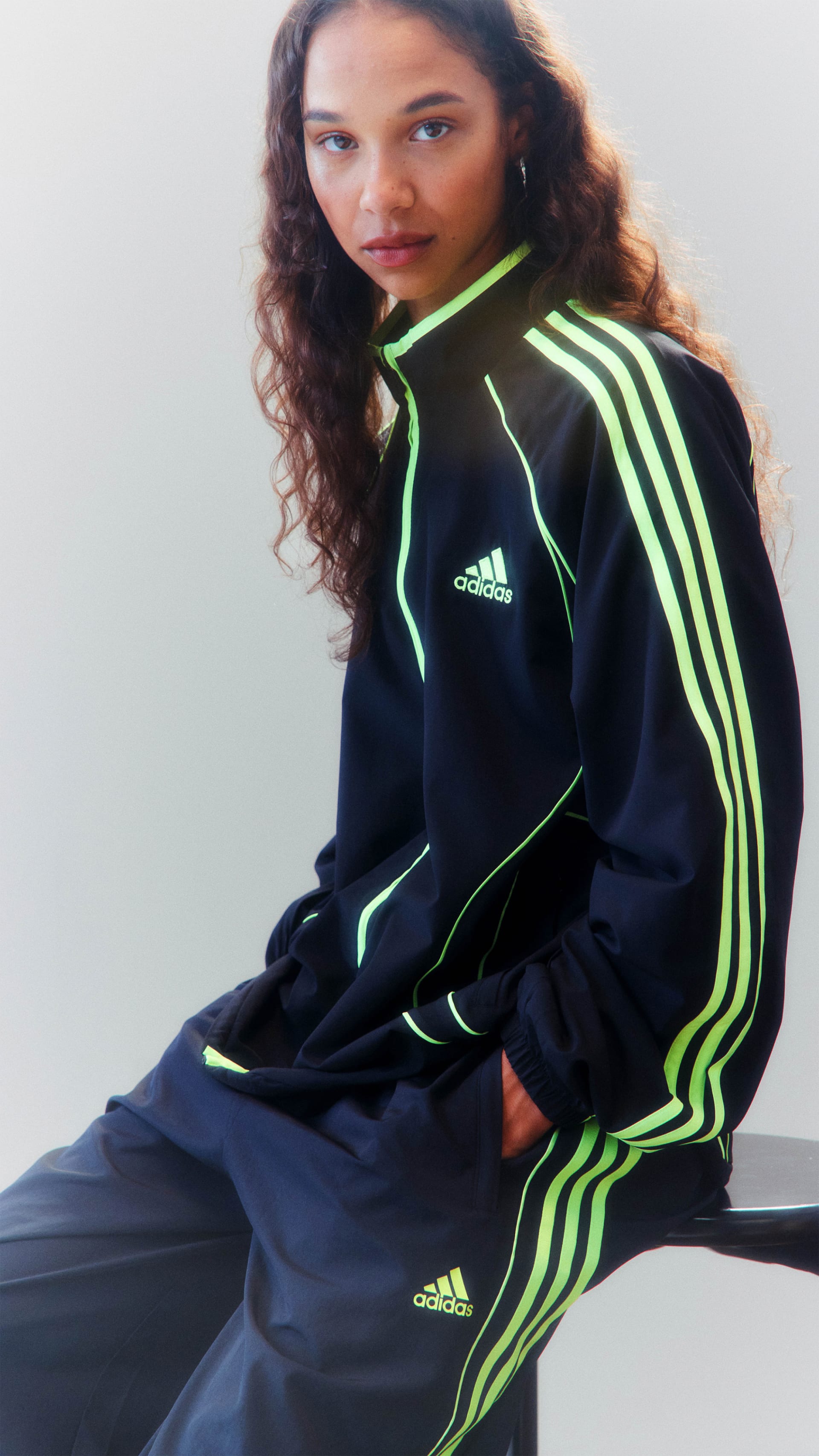 The image shows a woman wearing a black and neon green Adidas tracksuit, sitting on a plain background.