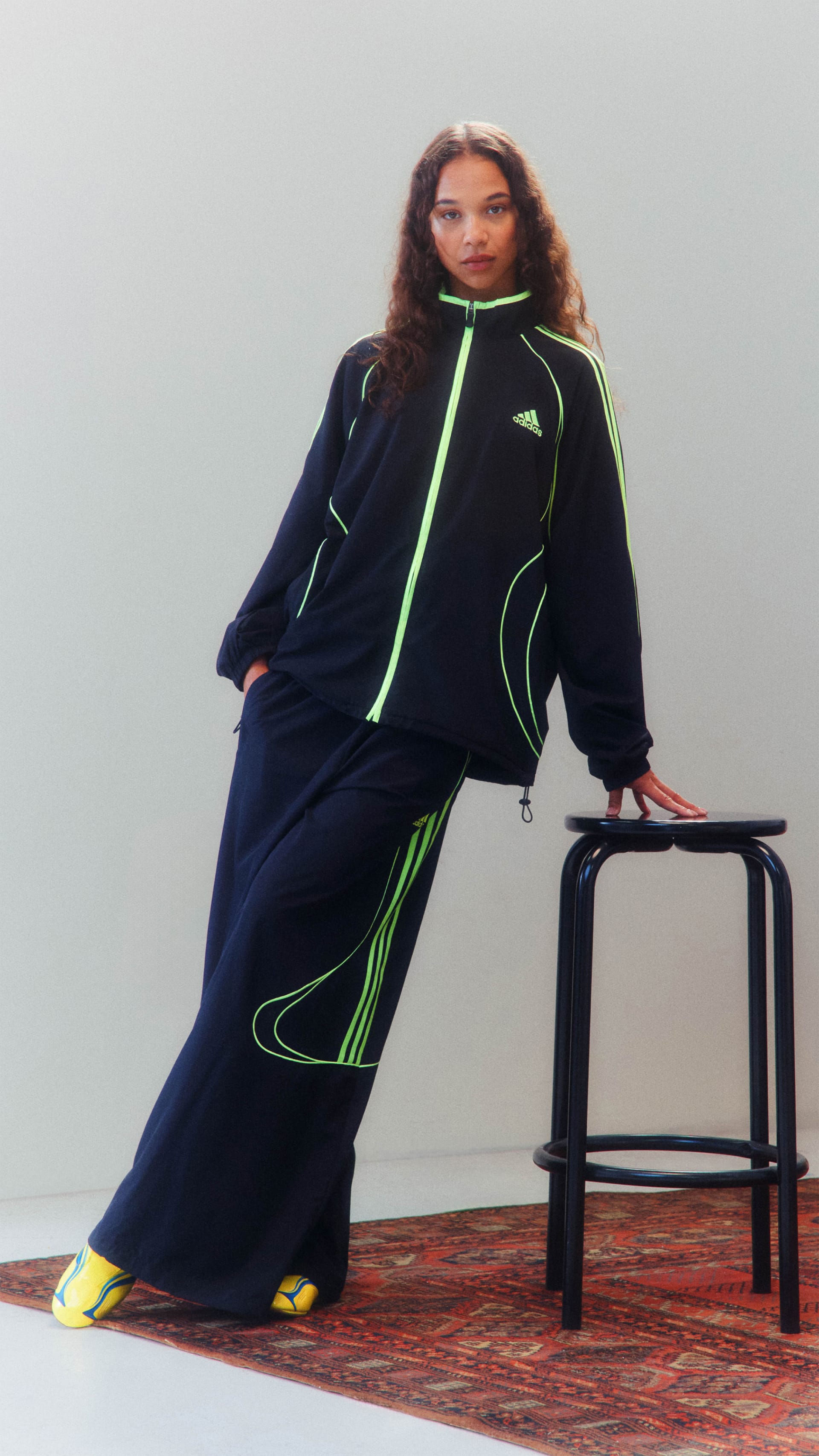 A person wearing a black and neon green tracksuit is sitting on a wooden stool against a plain background.