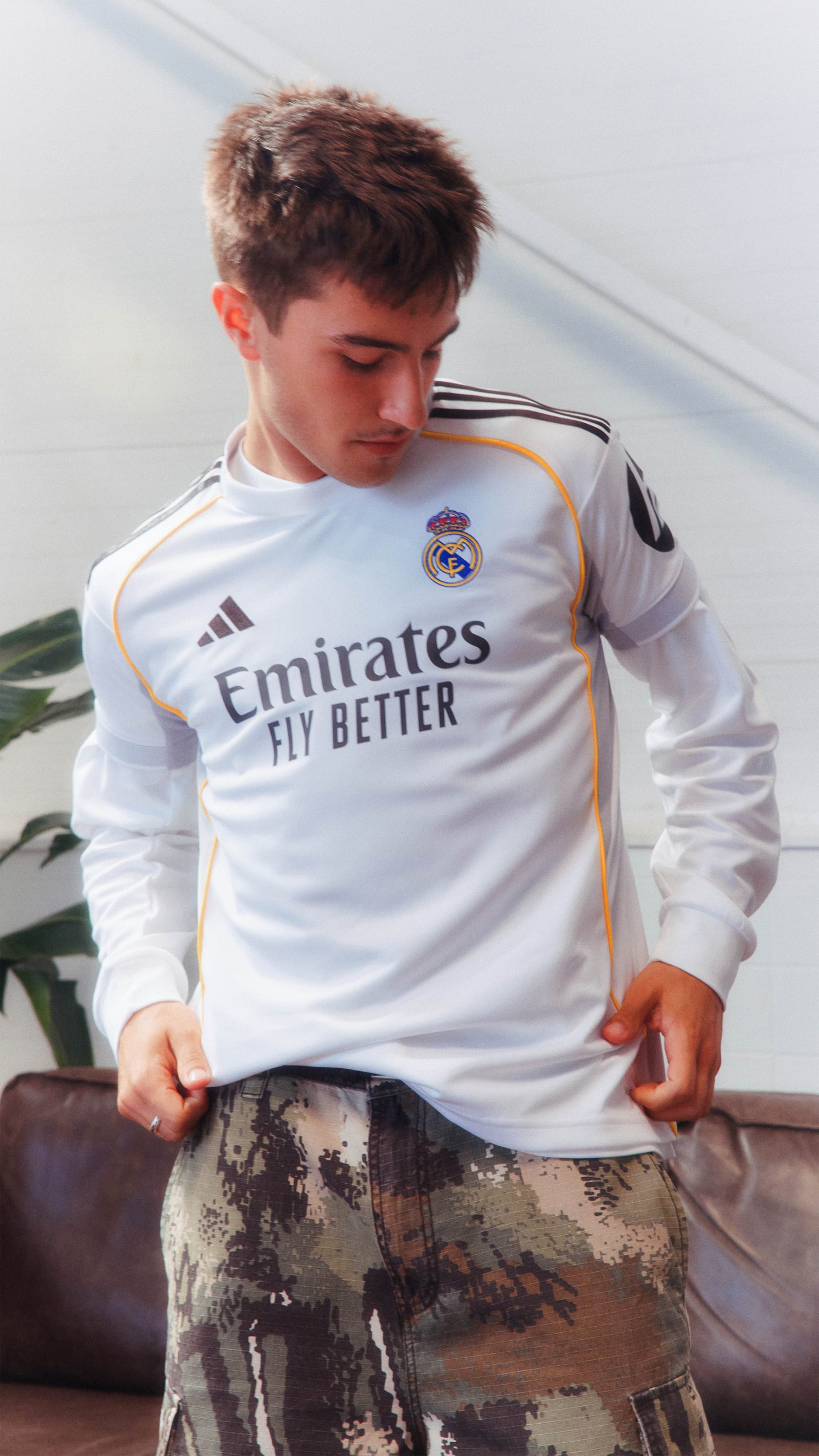 The image shows a young man wearing a white soccer jersey with the "Emirates" logo, sitting on a couch with a patterned background.