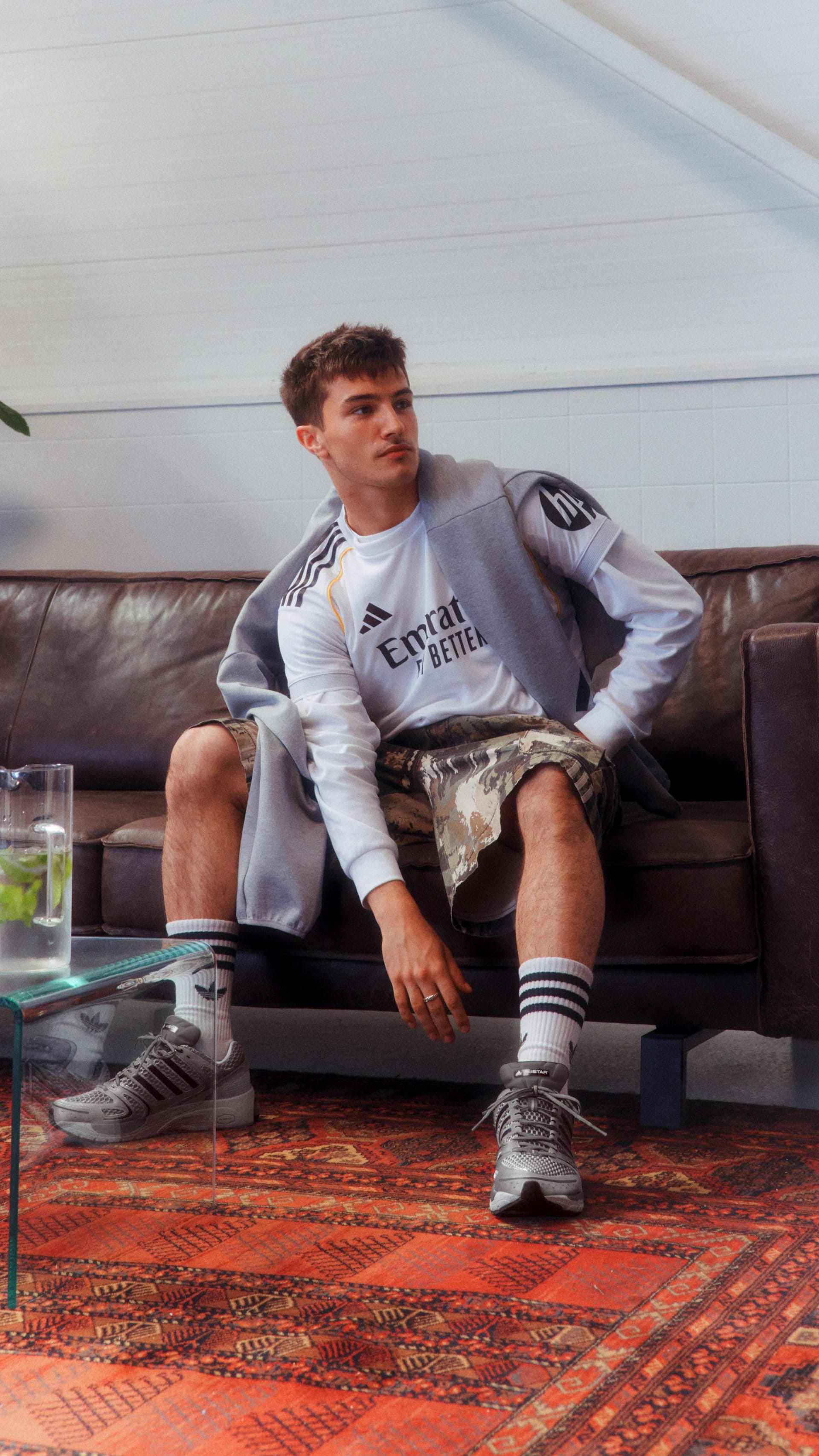 A young man wearing a gray sweatshirt and shorts is sitting on a couch, with a patterned rug and glass table visible in the background.